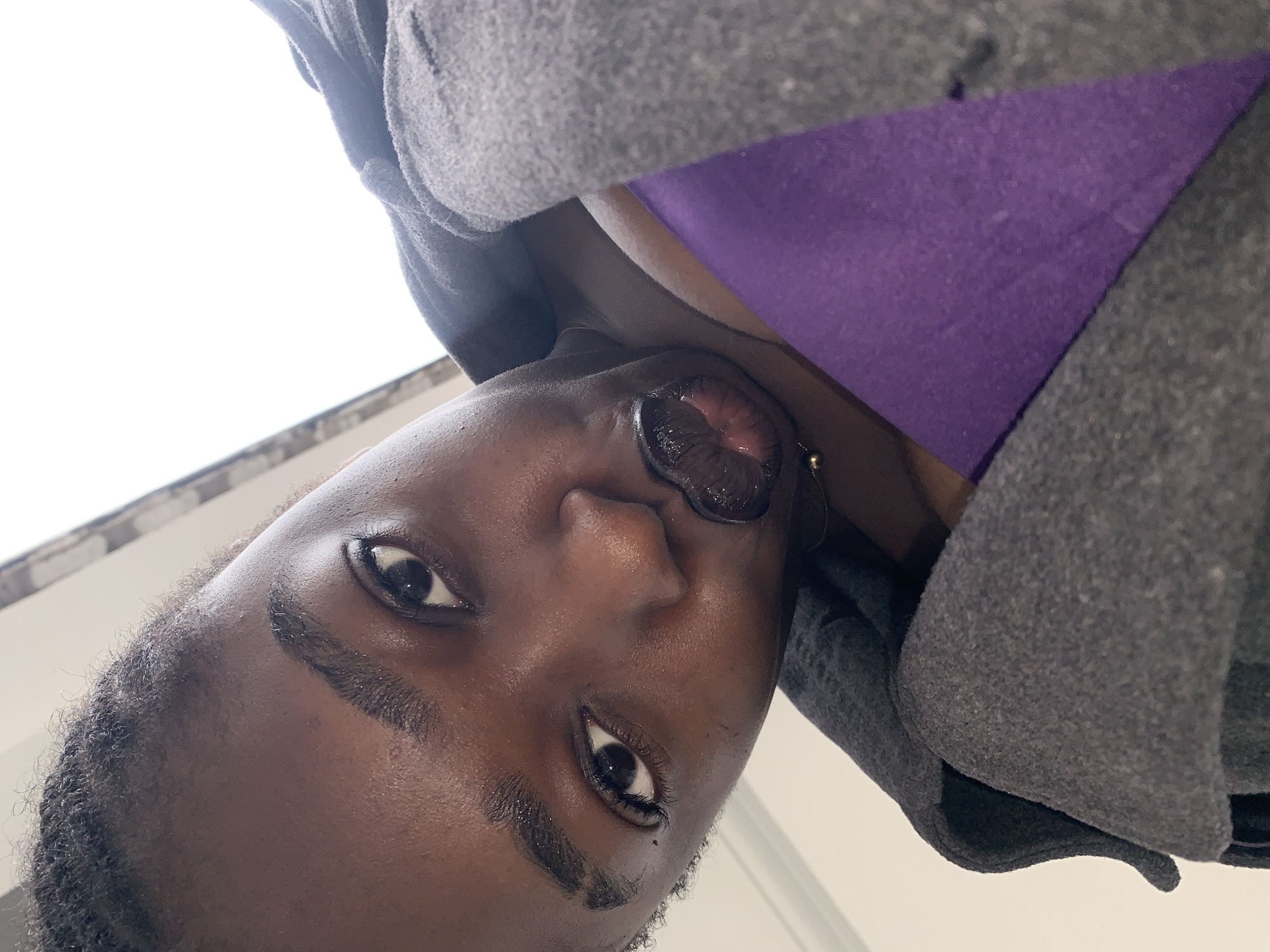 A close-up, high-angle shot shows a young Black woman making a kissing face towards the camera. She is wearing a dark grey jacket over a purple top. Her dark hair is pulled back. Her eyebrows are filled in, and she has on dark lipstick. A thin gold chain is visible around her neck. The background is out of focus, showing a bright, possibly outdoor, environment. The image appears to have been taken in Gura Top, Nigeria.