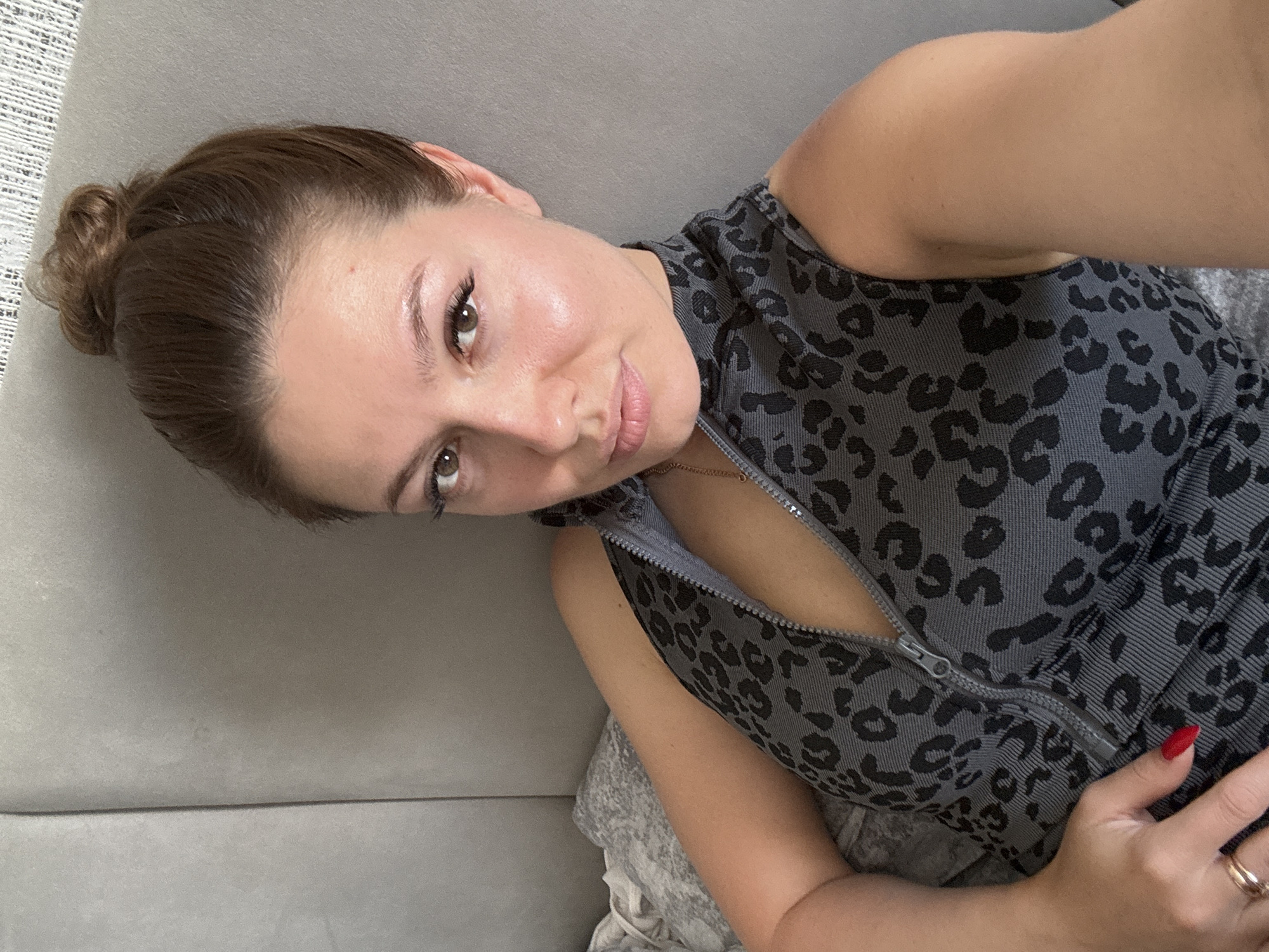 This is a close-up, angled selfie of a woman with brown hair pulled into a bun. She is lying on a grey couch, and the lighting suggests it is daytime. The woman is wearing a grey sleeveless top with a black leopard print pattern and a zipper down the front. Her skin appears dewy and radiant. She is looking at the camera with a gentle smile. Her right hand, with red nail polish, is resting on her chest, and she is wearing a ring on her ring finger. The background is a plain grey couch.