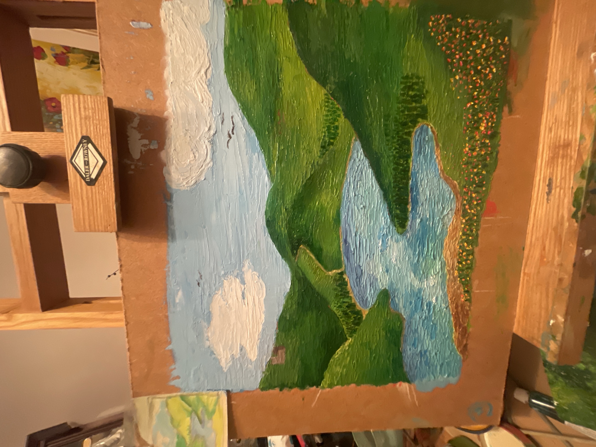 The image shows an oil painting of a landscape with a river running through green hills, set against a blue sky with white clouds. The painting is mounted on a wooden easel. The easel is branded with a “Daler-Rowney” label. There are no people visible in the image. The environment is indoors, presumably an artist's studio. There is no evidence to suggest the location is Maidstone, United Kingdom.