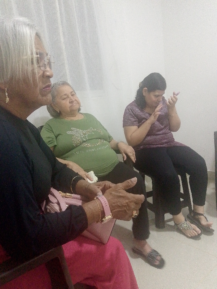 Three women are seated indoors. The woman in the foreground has gray hair, glasses, a black top, and a pink skirt, with a pink handbag visible. The woman in the middle wears an olive green shirt and black pants. The woman on the right, seated on a small stool, has curly hair, a patterned purple shirt, and black pants; she is focused on a phone in her hands. All three women appear to be elderly.