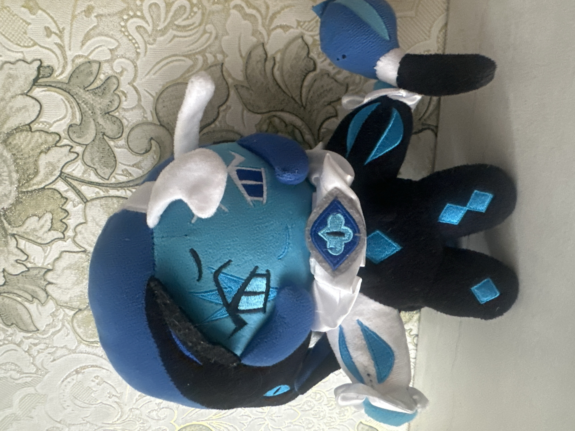 A plush toy resembling the character Frost Cookie from the game Cookie Run is positioned in a standing pose on a white surface, with a patterned wallpaper in the background. The plush toy is predominantly blue and black, with white accents. It has a large, round blue head with black accents around the sides and top. The face features closed eyes with blue markings and a small blue nose and mouth. White, horn-like protrusions extend from the top of its head. A white collar with a blue gem-like design adorns its neck. The body is black with blue diamond shapes. It has two arms; the left arm is partially visible and appears to have white and blue accents. The right arm is extended forward, holding a blue object with white and blue accents. The toy is facing slightly to the right. The wallpaper behind the toy has a light color with an ornate, floral pattern. The scene appears to be indoors, on a flat surface that could be a shelf or table. The lighting suggests it is daytime or well-lit indoors. There are no people or animals visible in the image.