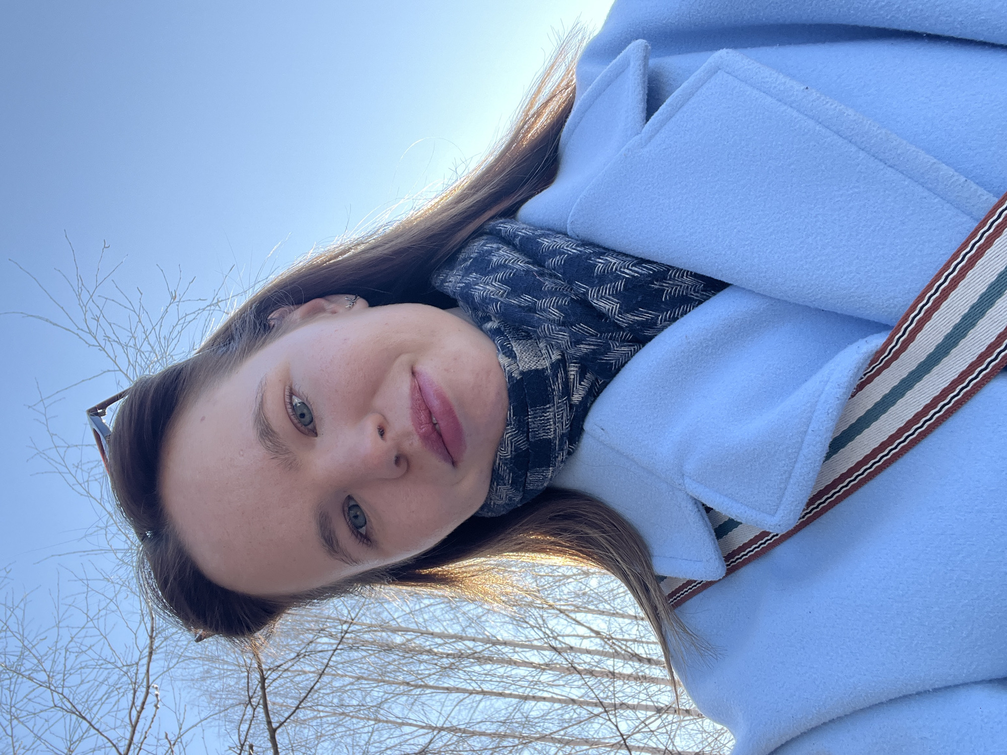 A female subject is depicted in an upward-angled, close-up portrait against a clear blue sky. She has light skin, blue eyes, and light brown hair with sunlit highlights. Dark-rimmed sunglasses are perched on her head. She wears a light blue overcoat with a wide collar and a dark scarf with a light chevron pattern around her neck. A striped strap, composed of red, white, green, and dark blue bands, crosses her chest, suggesting a bag strap. Small, silver-toned, antler-shaped earrings are visible in her left ear. The subject gazes directly at the camera with a slight, closed-mouth smile. The background consists of a bright, cloudless blue sky with numerous bare deciduous tree branches and slender trunks, consistent with birch trees, silhouetted against it. The lighting indicates a sunny outdoor environment. The specific location within Chaikovsky, Russia, cannot be determined from the visual information provided beyond an outdoor setting with trees.