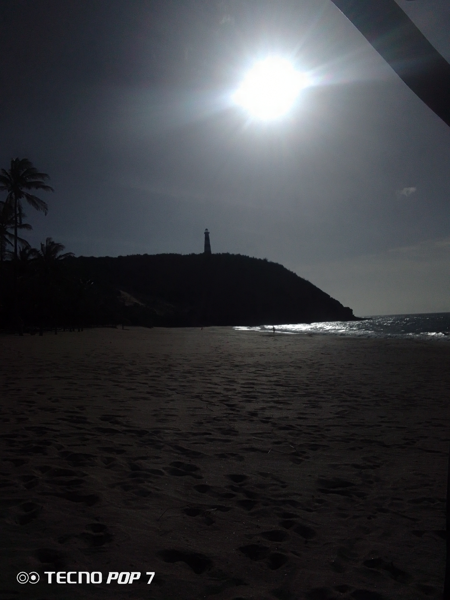 The image shows a beach scene in Pedro González, Venezuela. The bright sun dominates the sky, creating strong silhouettes. The beach is in the foreground. A dark, tree-covered hill rises in the middle ground, with a lighthouse silhouetted on top. The ocean is to the right, with visible waves. There are several people on the beach. The image is dark and underexposed. The words "TECNO POP 7" are in the bottom left corner of the image.