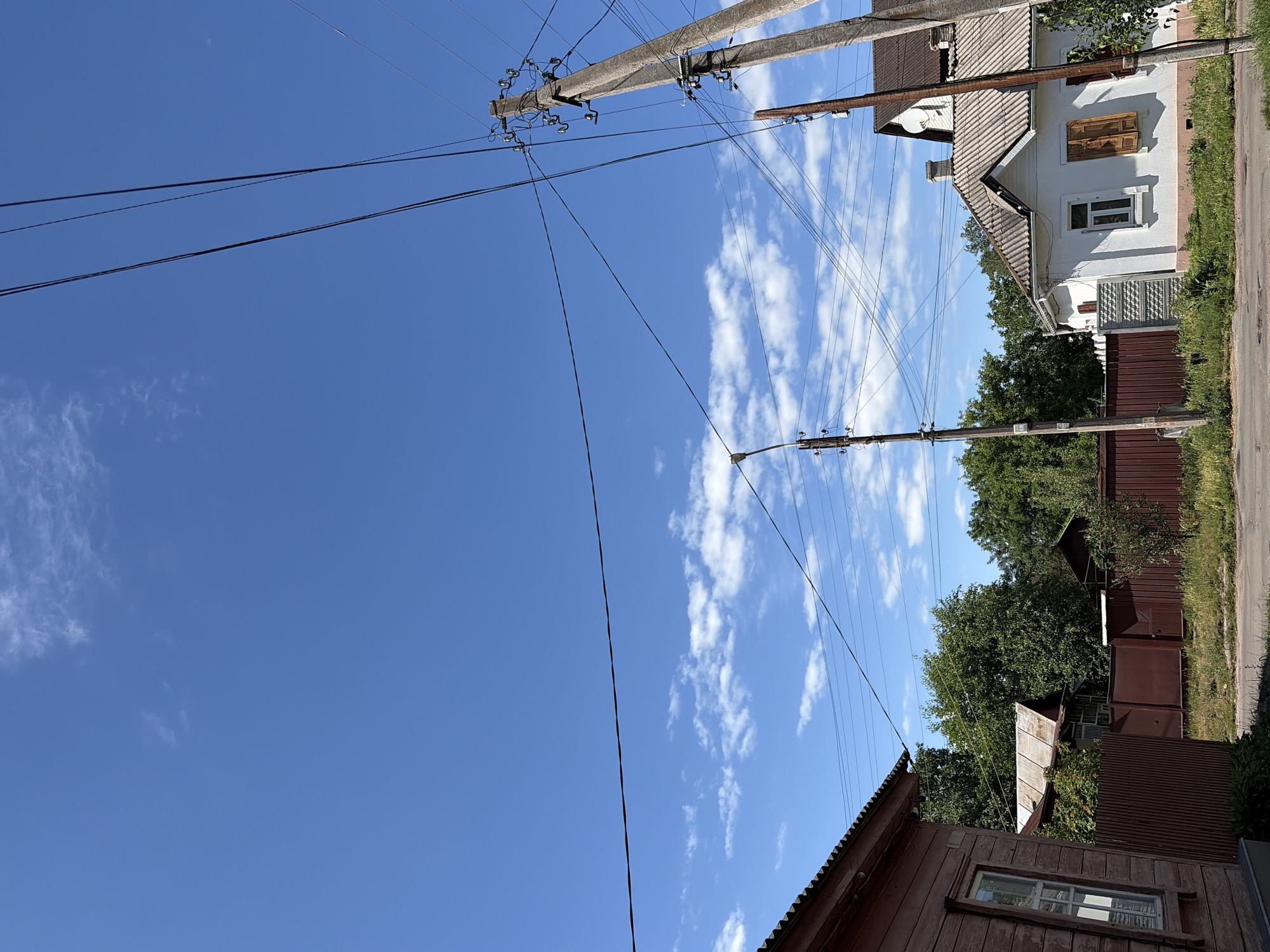 The image shows a street scene in Chernihiv, Ukraine, under a bright blue sky with scattered clouds. Several power lines crisscross the frame, supported by concrete utility poles. On the right side of the image, a white house with a grey tiled roof and wooden shutters is visible. Adjacent to it is a brown metal fence and lush green trees. On the left side, a portion of a wooden building with a corrugated metal roof can be seen, also bordered by a brown fence. The ground in the foreground appears to be a paved road with some patchy grass along the sides. No people are present in the image. The scene depicts a typical residential area in a town or city.