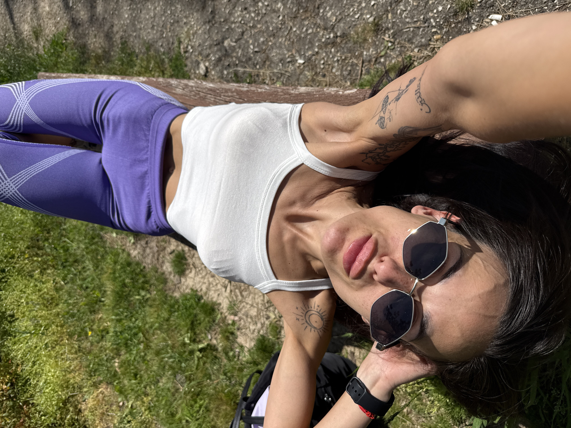 A woman lies outdoors, likely on a wooden bench, in what appears to be a park in Pleven, Bulgaria. She is seen from a high-angle perspective, possibly a selfie, looking upwards. She wears a white sleeveless tank top and purple leggings with a light-colored, geometric crisscross pattern. Her dark hair is spread out beneath her head.
She has several tattoos: one on her right upper arm features abstract, sketch-like designs, and another on her left shoulder/chest area depicts a stylized sun and crescent moon. She wears dark, hexagonal-framed sunglasses, partially pushed up, revealing her eyes. A black digital watch and a thin red bracelet are on her left wrist. Her lips are visible and appear slightly glossy.
The background shows patches of green grass interspersed with areas of dry earth or gravel, indicating a natural, unpaved outdoor environment. Bright sunlight casts strong shadows, suggesting it is midday or early afternoon on a clear, sunny day. The woman's relaxed posture and direct gaze towards the camera convey a sense of calm or leisure. No discernable text is visible in the image.