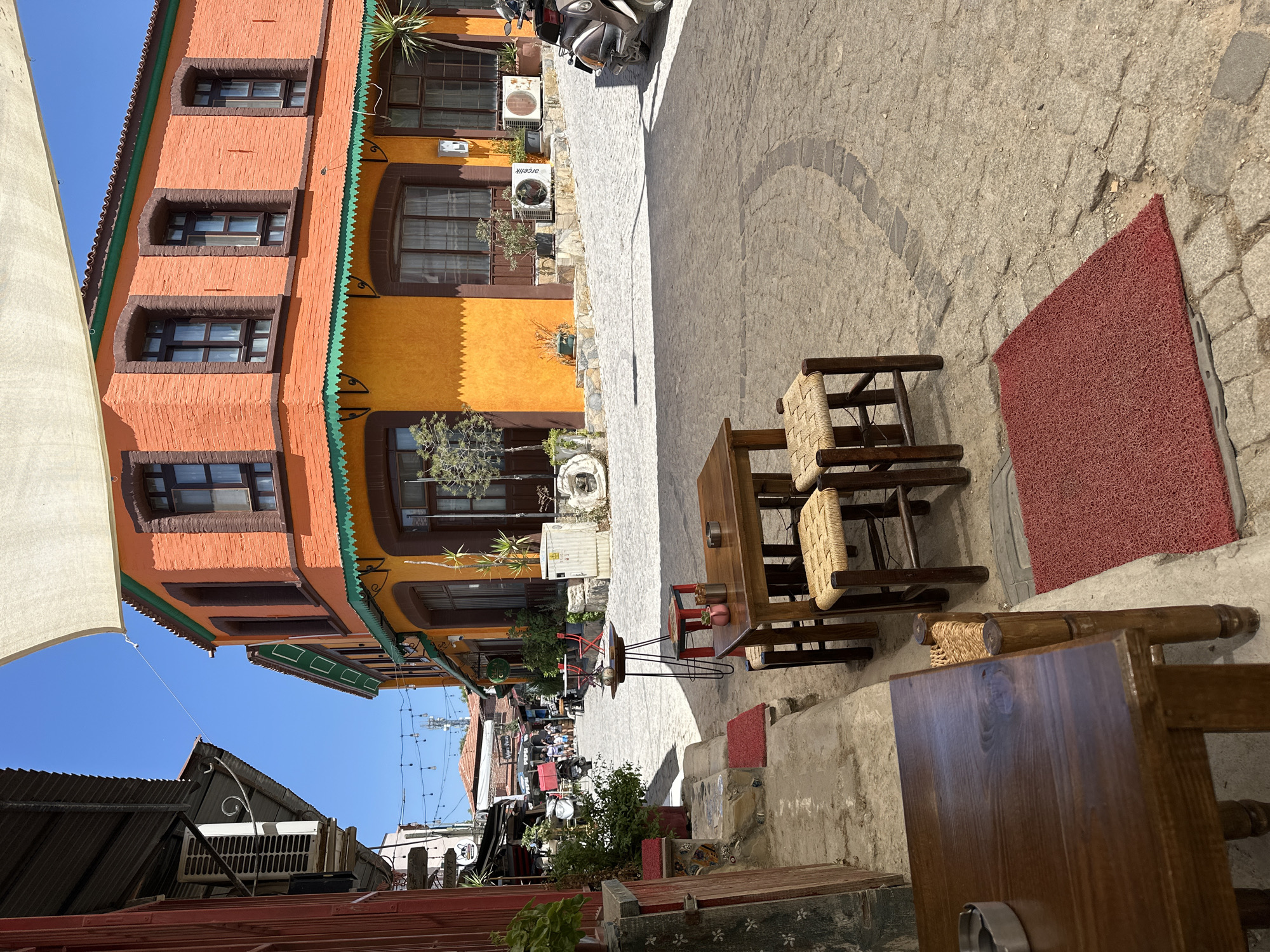 In the city of Urla, Turkey, on a sunny day, a quaint street scene unfolds with cobblestone paving. An orange and yellow building with a green trim corner dominates the right side of the frame, featuring arched windows. In the foreground, a wooden table and stools, typical of outdoor cafe seating, are set up on a red mat. The background reveals more of the narrow street, with shops and other buildings lining the sides. Several people are visible in the distance, walking and going about their day. A motorbike is parked near the orange building. The environment is characteristic of a Mediterranean town, with a relaxed and inviting atmosphere.