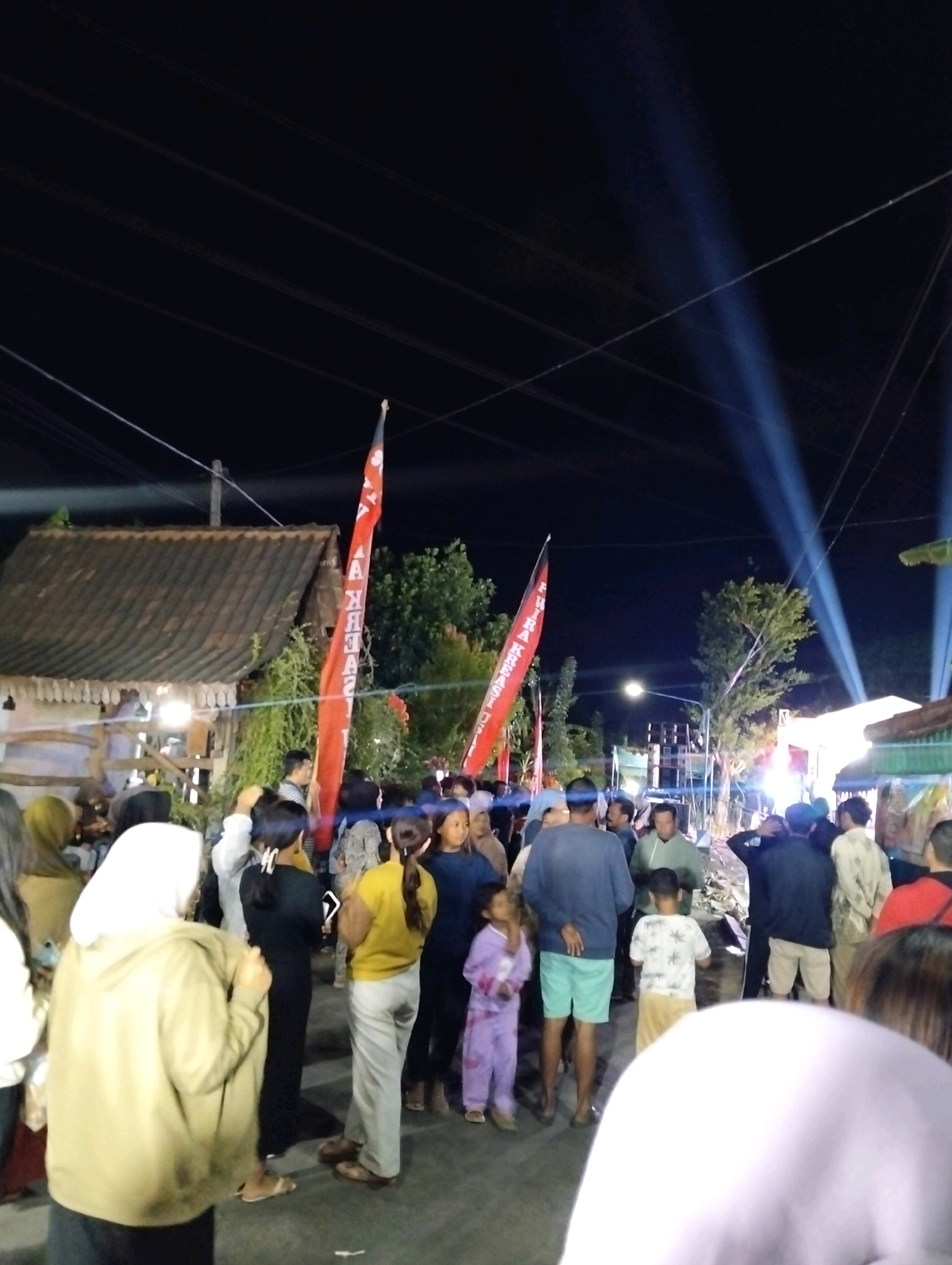 The image depicts a nighttime outdoor gathering in the city of Madusari, Indonesia. A crowd of people are congregated in what appears to be a street or open area. In the foreground, a person wearing a light brown hooded sweatshirt and a white hijab is partially visible from the side. To their left, a woman in a green hijab is also visible.
Further into the scene, several individuals are standing and interacting. A child in purple pajamas is looking to the left. A man in a blue long-sleeved shirt and turquoise shorts stands facing away. Another man in a light green shirt is facing the viewer. A group of people are gathered around what seems to be a stage or performance area, indicated by bright lights and speakers in the background.
Two prominent red flags with white text are visible, one on the left and another towards the center-right of the image, adding a festive or event-specific element to the scene. The background shows trees and some structures, including a building with a tiled roof on the left. Overhead, power lines are visible against the dark night sky, with visible light beams cutting through the darkness, suggesting artificial lighting for the event. The ground appears to be asphalt or a similar paved surface. The overall atmosphere suggests a lively community event or celebration.