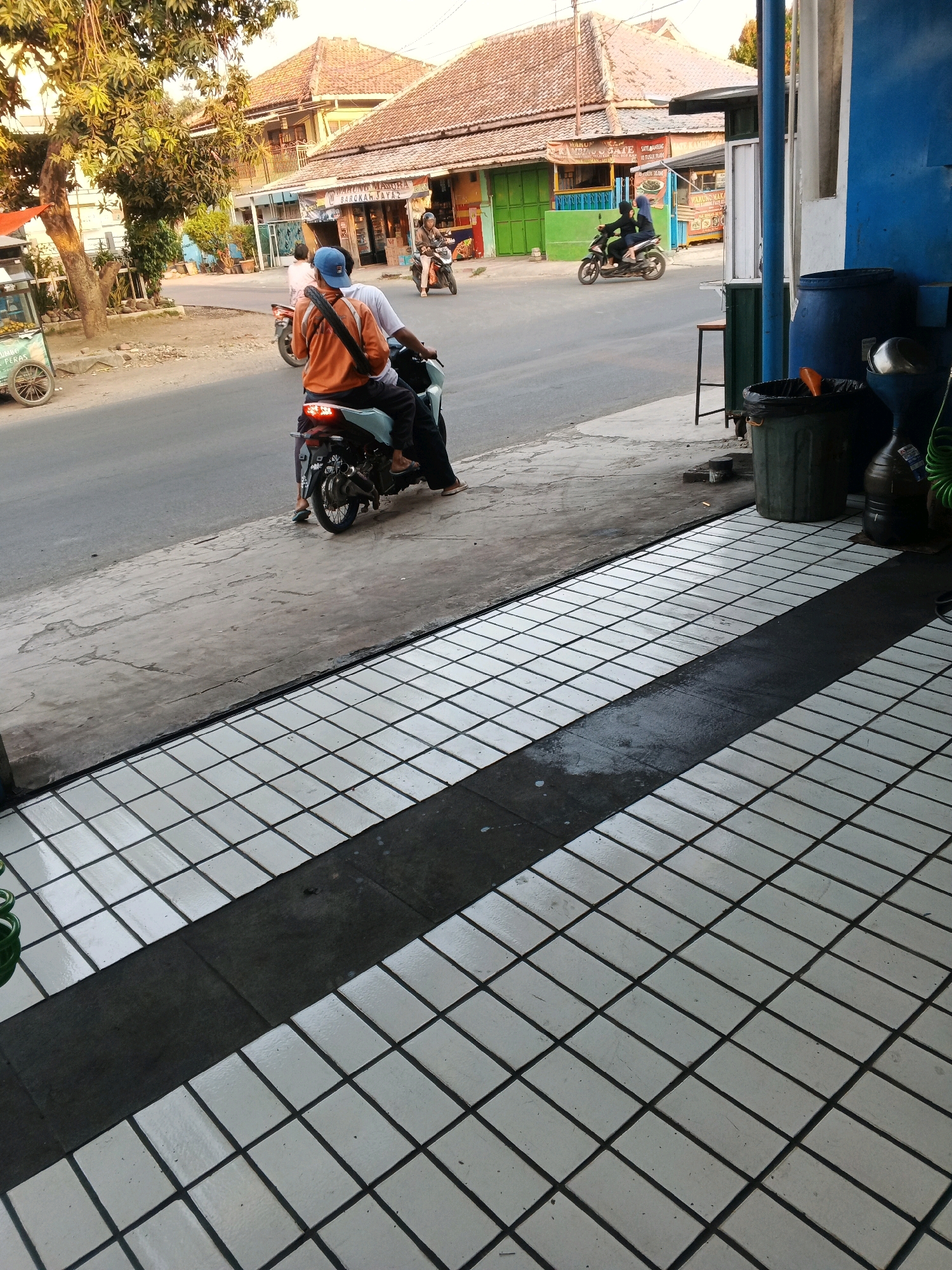 The image depicts a street scene in Taman, Indonesia, focusing on a tiled walkway in the foreground and a street with a few motorcycles and buildings in the background. The walkway is paved with white square tiles laid in a grid pattern, with a dark grey strip running diagonally across it.
On the street, two individuals are riding a motorcycle, with the driver wearing a blue cap and an orange jacket, and the passenger behind him. Another motorcycle with two occupants is seen further down the street. In the background, there are buildings with tiled roofs, typical of Indonesian architecture, and a few trees. The sky is visible in the upper part of the image, suggesting it might be late afternoon or early morning due to the warm light. Trash bins and a large blue barrel are visible near the buildings on the right side of the image, suggesting the area might be commercial or residential. The overall scene is an ordinary street view in a town in Indonesia.