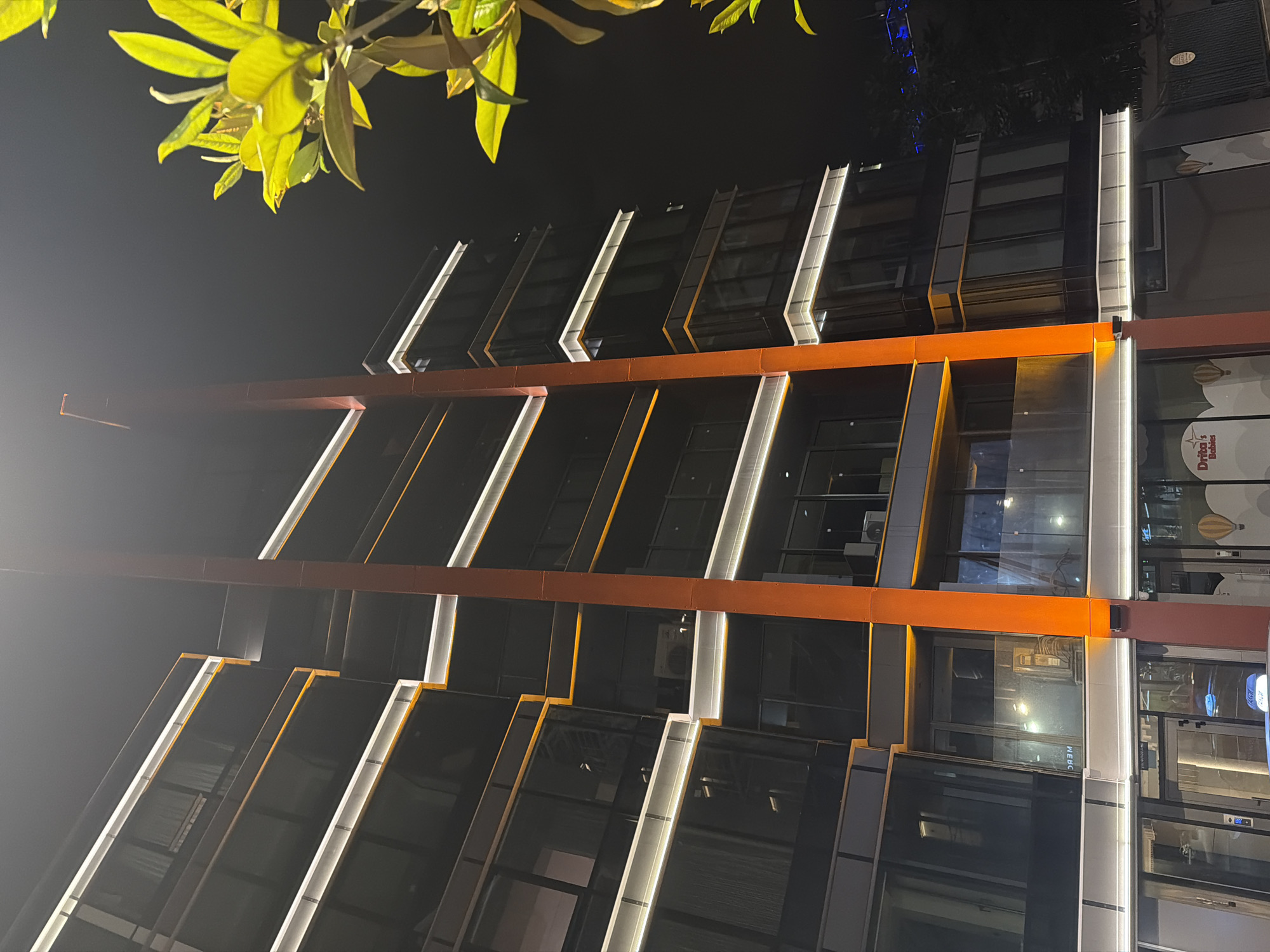 An upward-angled, nighttime photograph captures the illuminated facade of a modern multi-story building in Sofia, Bulgaria. The sky is dark, indicating night. The building features dark glass windows and a prominent architectural design with alternating sections.
Bright, cool-white LED strip lights run vertically along the edges of many building sections, creating strong visual lines against the dark facade. A distinctive horizontal orange beam cuts across the building's mid-section, providing a warm contrasting color and intersecting the vertical lights. The building appears to be commercial or mixed-use.
In the upper left corner, vibrant green leaves of a tree are visible against the dark background, lit by an unseen light source. On the far right side of the building, a sign is partially visible with the text "Draba's Babies," suggesting a children's clothing store or similar business. This sign also features decorative elements resembling hot air balloons. Reflections are visible in some windows, including what appears to be an air conditioning unit on an upper floor and the partial word "WEBC" or similar in a lower window. There is no explicit activity or specific event taking place, just the static scene of an illuminated urban structure. The overall impression is one of modern architecture.