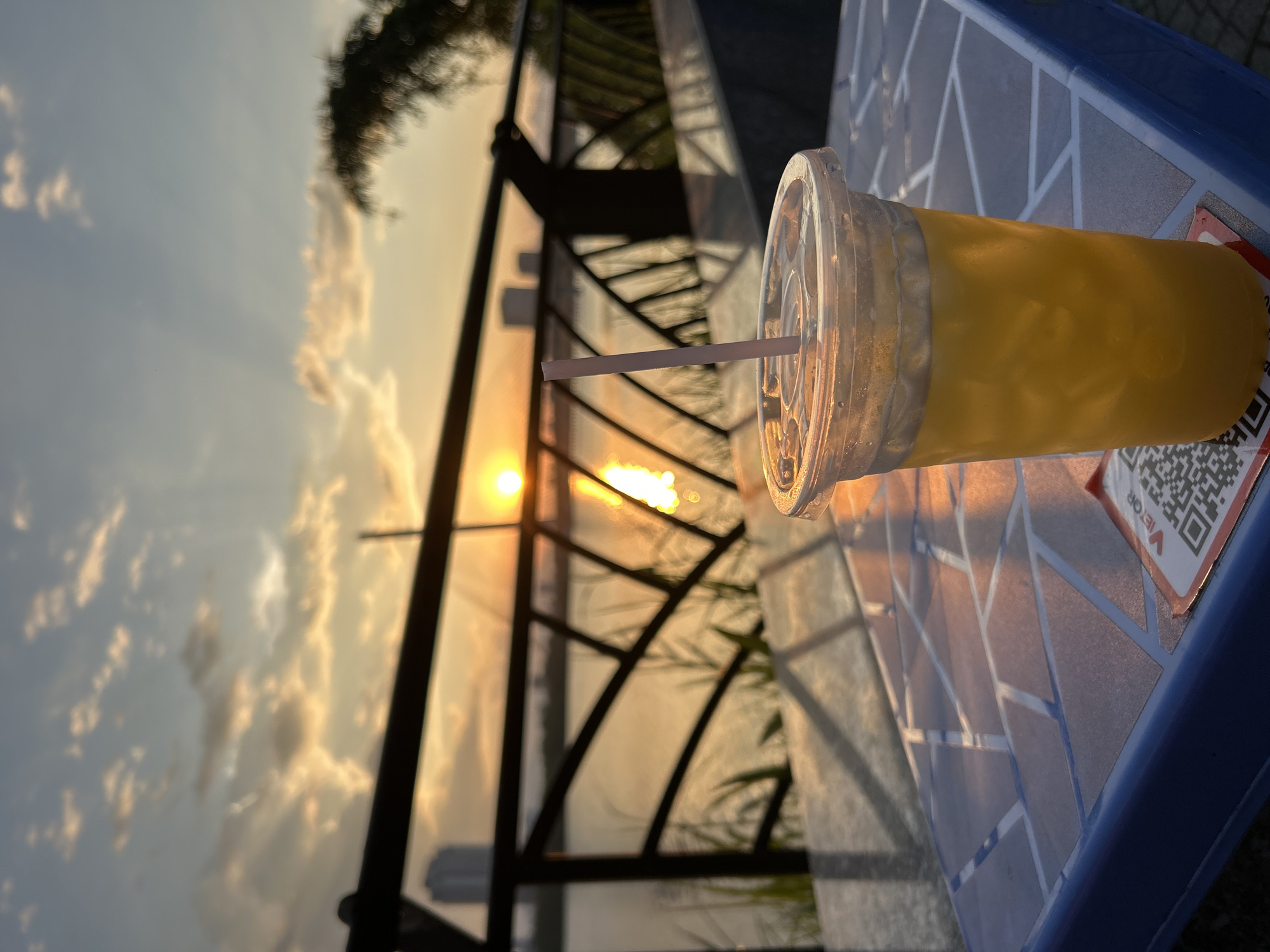 An outdoor table with a blue tiled surface holds a clear plastic cup of yellow liquid with ice and a pink straw. The cup is positioned to the right of the frame. In the background, a body of water reflects the setting sun, which is a bright orange orb low in the sky. Clouds are scattered across the sky, illuminated by the sun's golden rays. A black railing runs horizontally across the middle ground, with tall buildings visible in the distance on the far side of the water. The scene appears to be a waterfront cafe or restaurant in the city of Đà Nẵng, Vietnam, likely at sunset. The overall atmosphere is serene and picturesque.