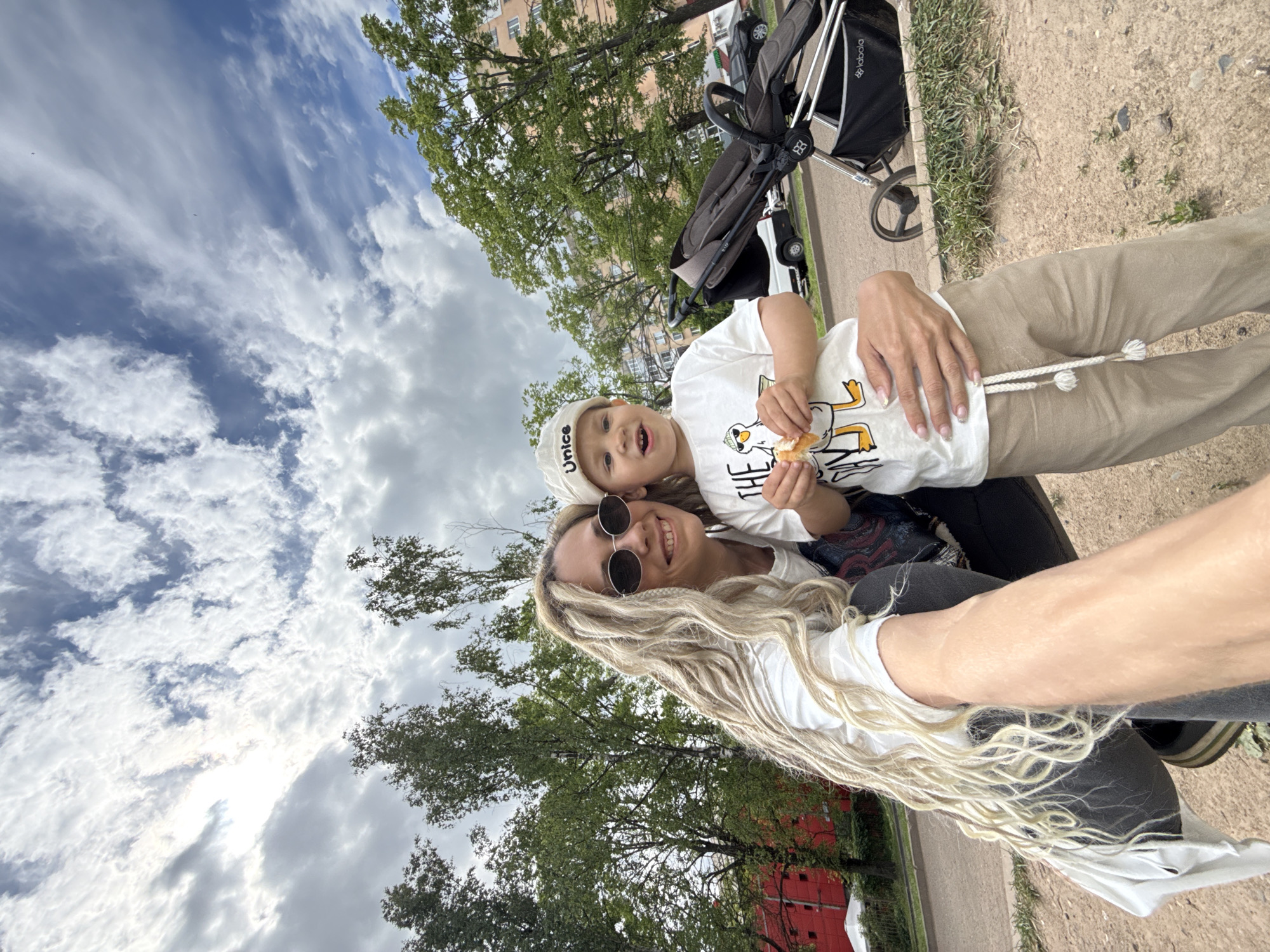 A brightly lit, outdoor portrait captures a woman and a young child, likely a mother and son, smiling happily for the camera. The image is taken from a slightly low angle, showing a broad expanse of cloudy blue sky above.
The woman is on the left, with long, blonde dreadlocks or thin braids cascading over her shoulder. She wears dark, round sunglasses and a white t-shirt, her arm reaching out towards the camera, suggesting a selfie. She has manicured nails and her hand rests gently on the child. Her smile is broad and joyful.
The child, positioned in front of the woman, appears to be held or sitting close to her. They wear a light-colored baseball cap with "Unice" written on the front and a white t-shirt featuring a cartoon duck graphic with partial text "THE". The child is holding a piece of bread or pastry and looks towards the camera with an open mouth, conveying excitement or a playful expression.
The setting is an outdoor area, possibly a park or a paved path next to a grassy section. In the background, lush green trees are visible, along with glimpses of a building, possibly an apartment complex. A black and grey baby stroller with the "lbabala" logo is parked nearby on the right. The ground beneath them is a mix of dirt and gravel, with patches of grass along the edge. The weather appears to be overcast but bright, indicating daytime. Both subjects exude a sense of warmth and happiness.