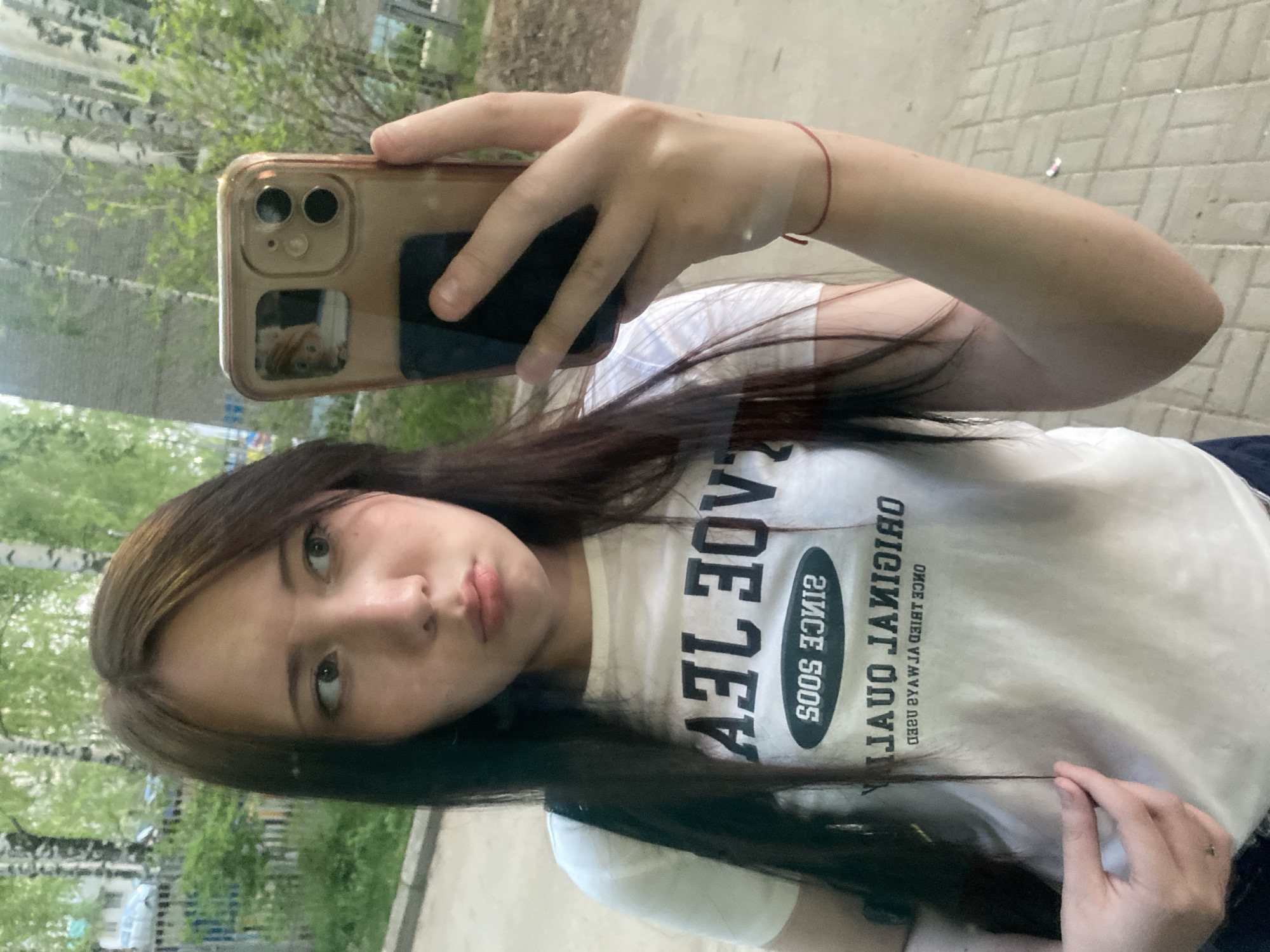 A young woman is taking a selfie in an outdoor setting. She is holding a smartphone with a gold-colored case, which is partially covering her face. Her hair is dark and falls across her chest. She is wearing a white t-shirt with a graphic print. The background shows trees and a building. The location appears to be an urban area.