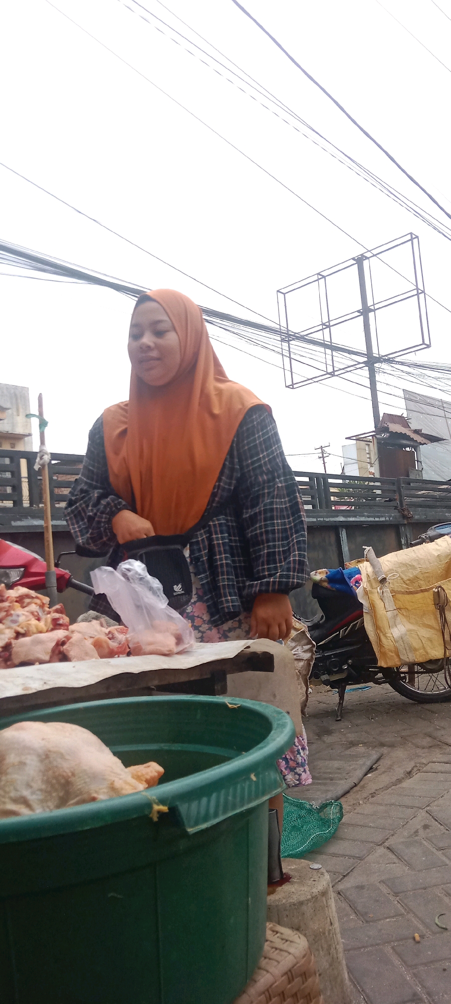 A young woman wearing an orange hijab and a plaid shirt is standing behind a market stall in Kras, Indonesia. She is holding a black fanny pack. In front of her on the stall are pieces of raw chicken, some wrapped in plastic. A large green tub containing a whole raw chicken is to her left. A motorcycle with a yellow tarp is parked to her right. The background shows a street with power lines and buildings.