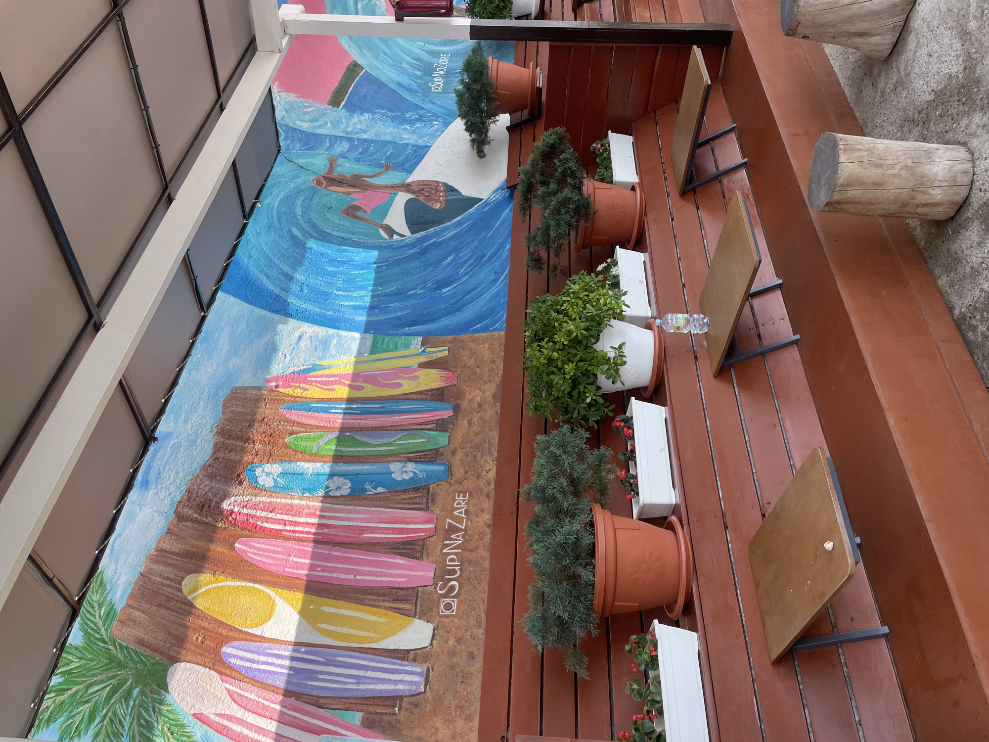 This image captures an outdoor seating area with a vibrant mural on the wall, located in Sochi, Russia. The mural depicts a beach scene with colorful surfboards lined up against a wooden fence on the left, and a surfer riding a wave on the right, against a bright blue and pink background. The text "SupNazare" is visible on the mural.
In front of the mural, a wooden bench runs horizontally, adorned with several potted plants. These include evergreen shrubs in terracotta pots and smaller plants in white rectangular planters, some with red flowers. Small, wooden fold-out tables are attached to the wall above the bench, with one table holding a plastic water bottle.
The seating area consists of tiered wooden benches painted a reddish-brown color, with the lower benches designed to hold planters or provide additional seating. Two rustic log stools are placed on the concrete ground in the foreground. The area is covered by a light-colored awning supported by a white structure, suggesting it's a sheltered outdoor space, possibly part of a cafe or restaurant. The overall impression is a relaxed, summery ambiance.