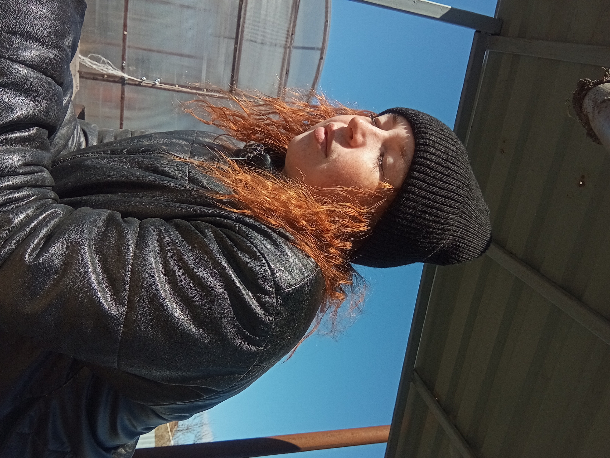 The image shows a person with red hair wearing a black beanie and black leather jacket, with their eyes closed, presumably outdoors under a clear blue sky. The location is identified as Yurgamysh, Russia. Overhead, there appears to be a corrugated metal structure and what might be a greenhouse roof. A pipe-like object is seen in the upper right corner, possibly part of the surrounding infrastructure.
