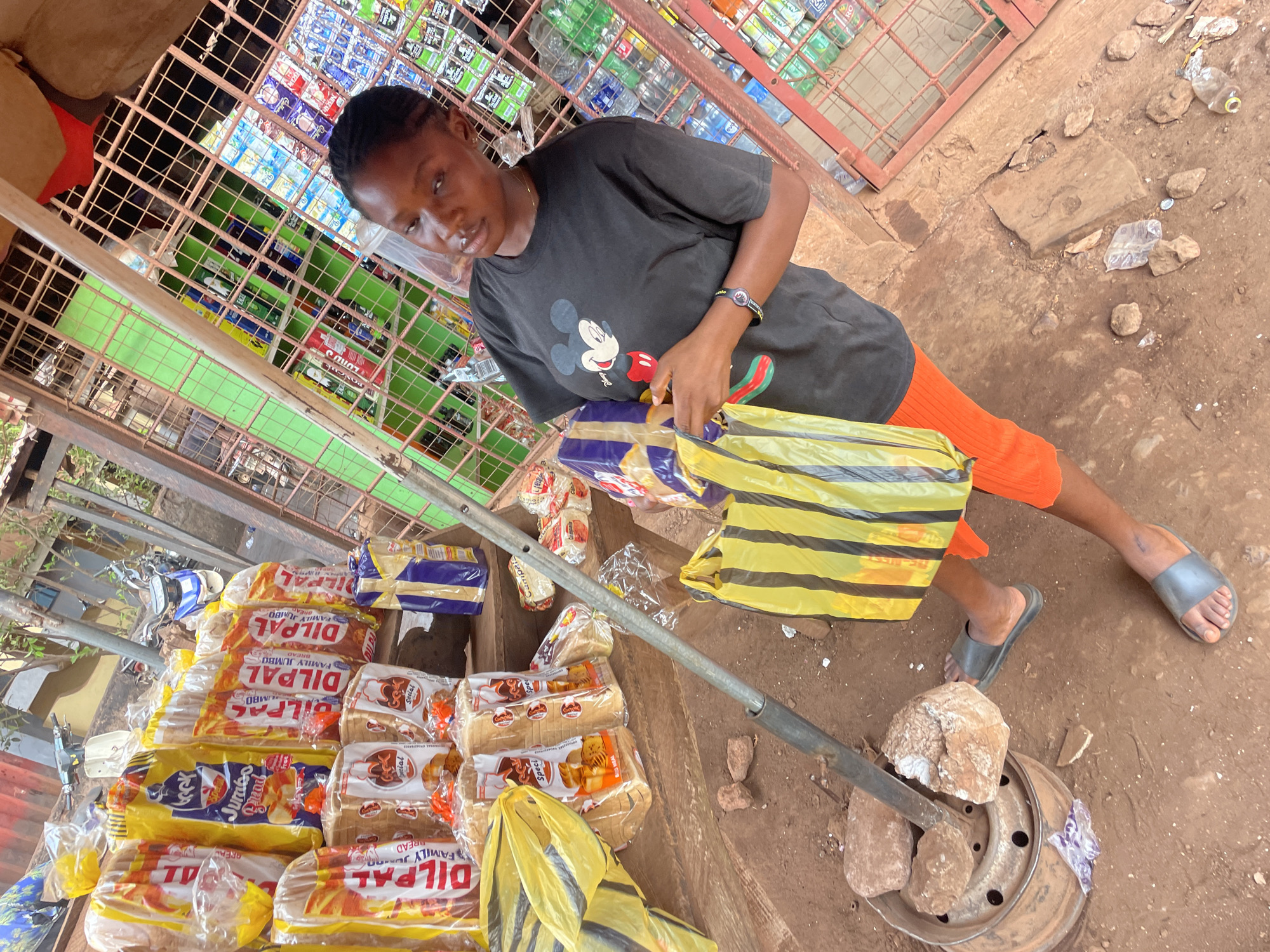 A woman stands near a roadside stall in Otukpa, Nigeria, packaging bread. The stall is a simple metal-framed structure filled with various goods. The woman wears a dark t-shirt with a Mickey Mouse graphic and orange shorts, and she is packing loaves of DILPAL brand bread into a yellow and black striped bag. The ground is dusty and scattered with debris. The stall is stocked with an assortment of items, including bottled drinks and packaged goods. A metal pole rests on the ground, secured by a metal base and rocks.