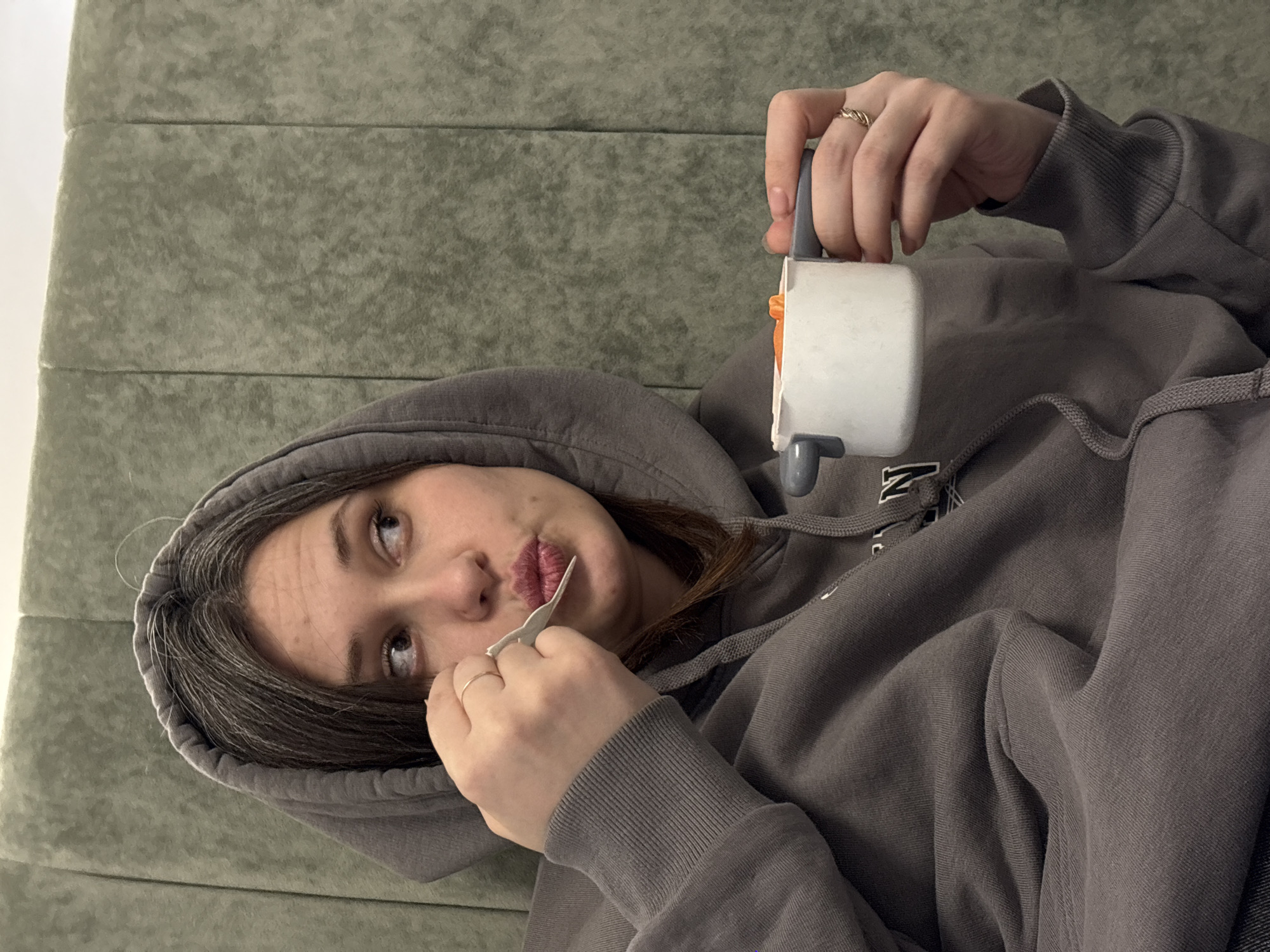 A woman in a grey hooded sweatshirt lies on a green upholstered surface, holding a lint roller in her right hand. She is holding a small piece of paper or film up to her face with her left hand. She is looking up and to the left. Based on the image content alone, it is not possible to confirm the location in Chaikovsky, Russia.