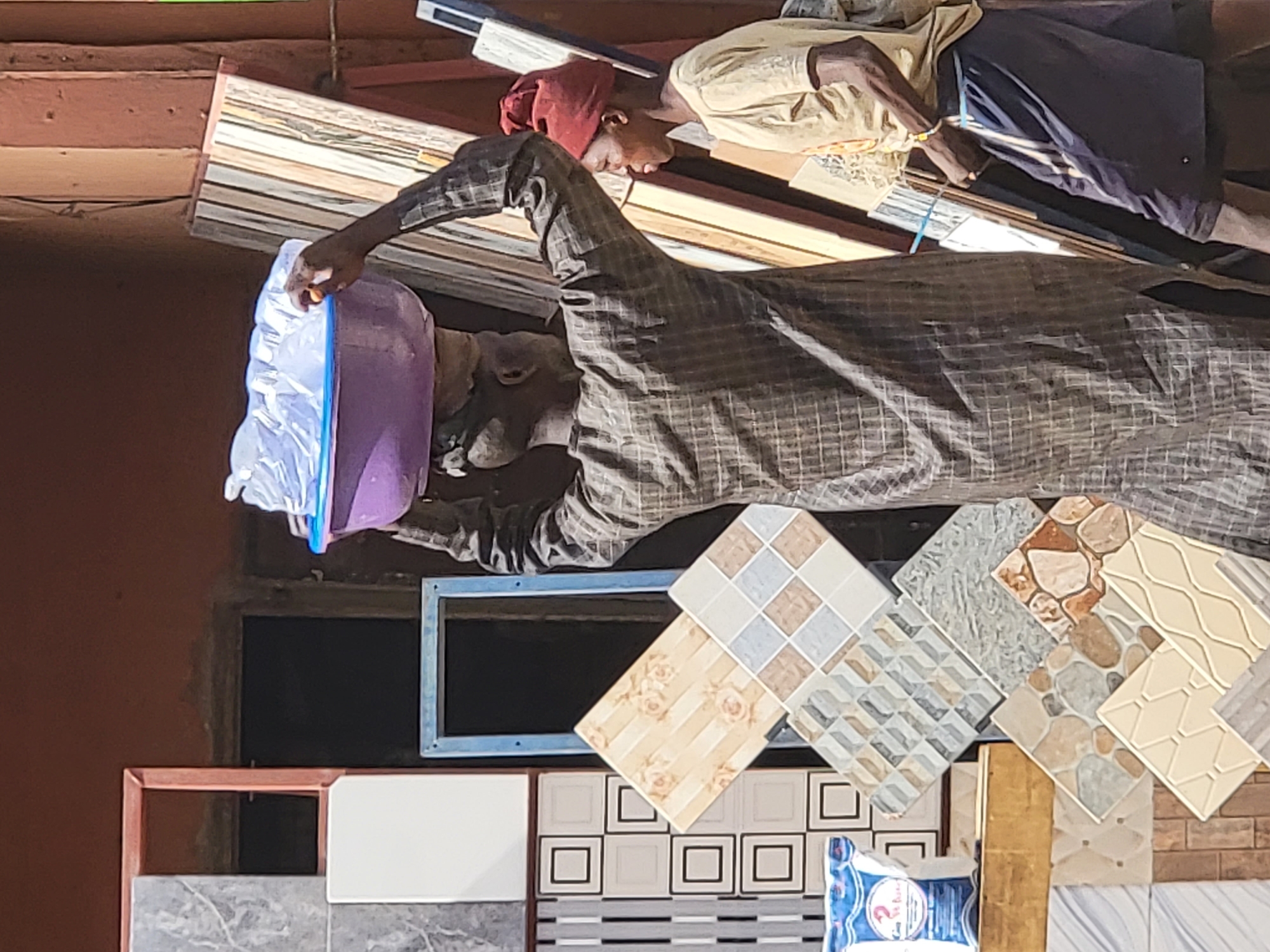 In this image, which appears to be rotated 90 degrees counter-clockwise but will be described as if upright, a vibrant commercial scene unfolds, likely a market stall specializing in building materials in Maiburiji, Nigeria.
Two individuals are present in the frame. In the foreground, a person wearing a plaid long-sleeved shirt is seen interacting with a large, purple container that is encased in a clear plastic bag. Their head is largely obscured, possibly by the plastic or a head covering. Behind them, a second individual, dressed in a light-colored long-sleeved shirt, dark pants, and a reddish head covering, stands near some wooden planks or shelving units, suggesting they are attending the stall.
The scene is dominated by an extensive display of various ceramic and porcelain tile samples, particularly in the lower and right sections of the image. These tiles showcase a wide array of patterns, including geometric squares, intricate floral designs, textured stone-like finishes, and contemporary wavy motifs, indicating a diverse selection for sale.
Other visible objects include wooden structures forming shelves or display racks, and what appears to be a solar panel or outdoor light fixture attached to an overhead beam in the background. A light blue-framed opening or signage is visible in the middle-left of the image. At the bottom, a package with a blue and white label is partially visible, with faint, unreadable text, possibly including "2 Brothers" along with other characters.
The bright lighting suggests it is daytime, with no specific weather conditions evident beyond clear skies. The overall activity centers around the display and potential sale of building materials, characteristic of a bustling market environment.