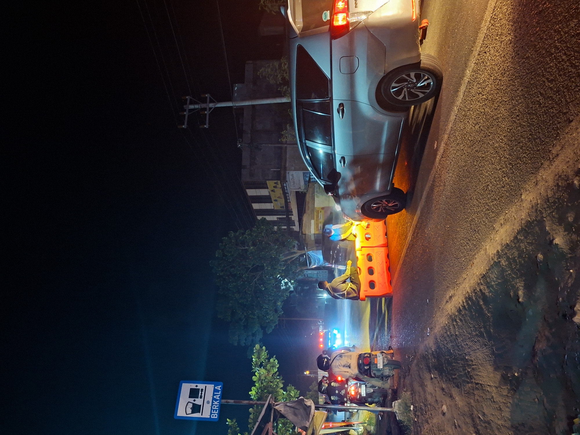 A silver Toyota Innova is parked on the side of a road in Lhokseumawe, Indonesia, illuminated by streetlights and the vehicle's taillights. The road has a rough, asphalt surface with dark, wet patches on the right side. Orange plastic barriers are positioned behind the car. A person in a light-colored shirt is sitting behind the barrier. Two motorcycles are parked nearby, with license plates visible. A blue sign reading "BERKALA" in white text with a graphic is on a post to the left. Background buildings and streetlights are visible in the distance. The sky is dark, suggesting it is nighttime.
