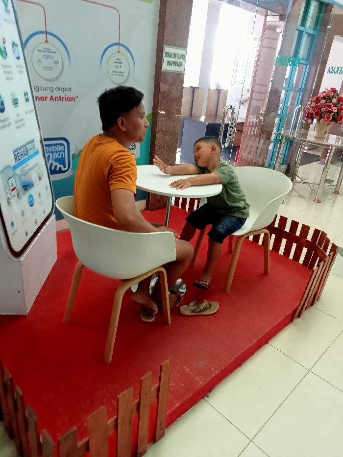 The image shows a man and a young boy sitting at a table in a shopping mall. The man is wearing a yellow shirt and is sitting on a white chair with wooden legs. The boy is wearing a green shirt and blue jeans. He is sitting on a white chair with wooden legs. They are sitting at a round, white table. The table is set on a red carpet surrounded by a small fence made of brown wooden planks. The man is looking at the boy, who is looking at the camera. The man has his feet flat on the red carpet. The boy is wearing sandals and his feet are off the ground, hanging from the chair. There is a large white sign with text in Indonesian and a small sign with a mobile phone. Behind the man and boy, there is a glass wall. In the distance, there is a potted plant and a chair. The location is the Kecamatan Margorejo area of Surabaya, Indonesia, at latitude: -6.76, longitude: 111.015.