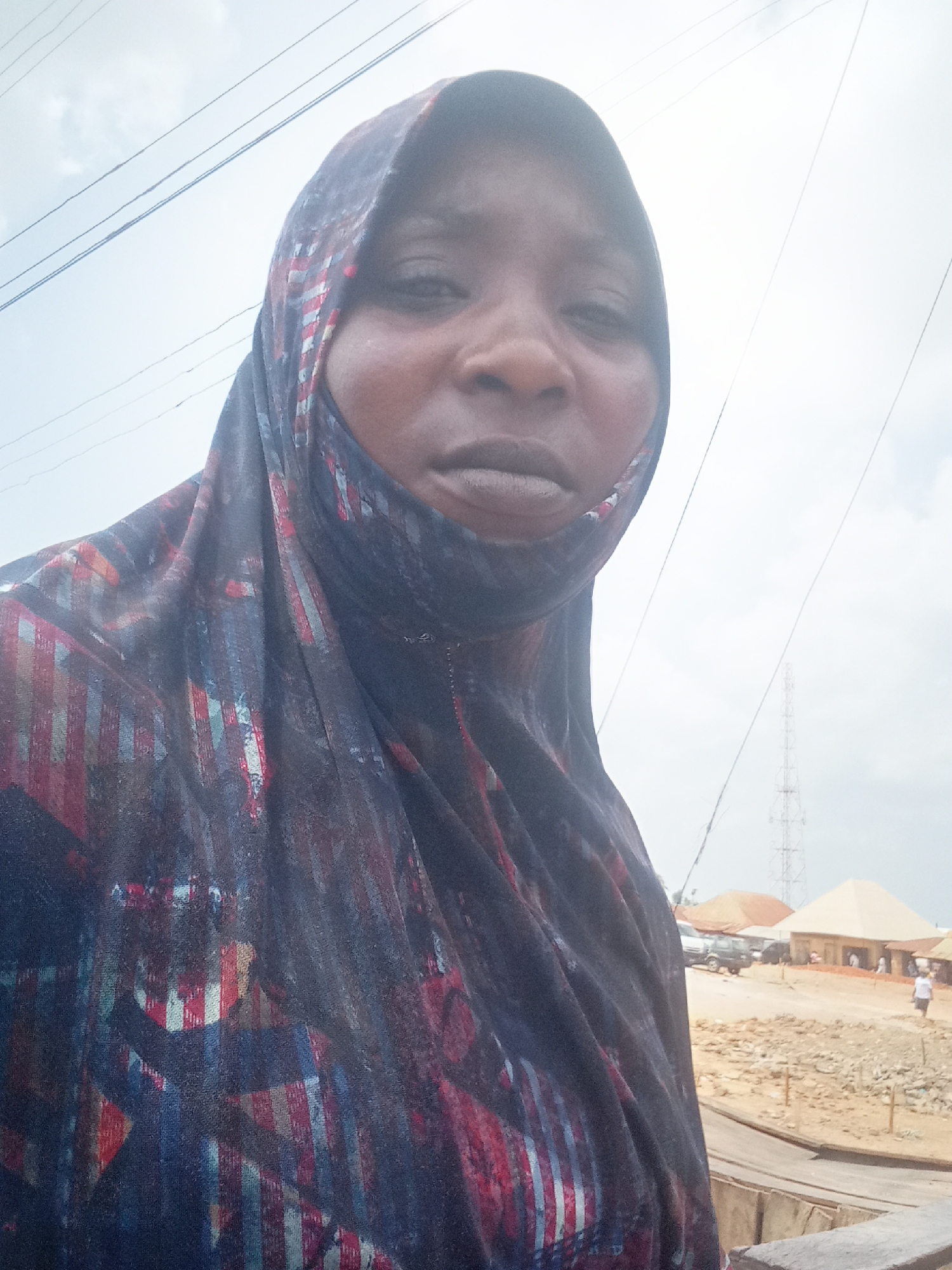 A medium shot captures a woman wearing a dark, patterned hijab. The backdrop reveals an outdoor scene in Akwanga, Nigeria. Visible are a dirt road, simple buildings, a telecommunications tower, a vehicle, and a person walking in the distance. Overhead power lines are strung against a partly cloudy sky.