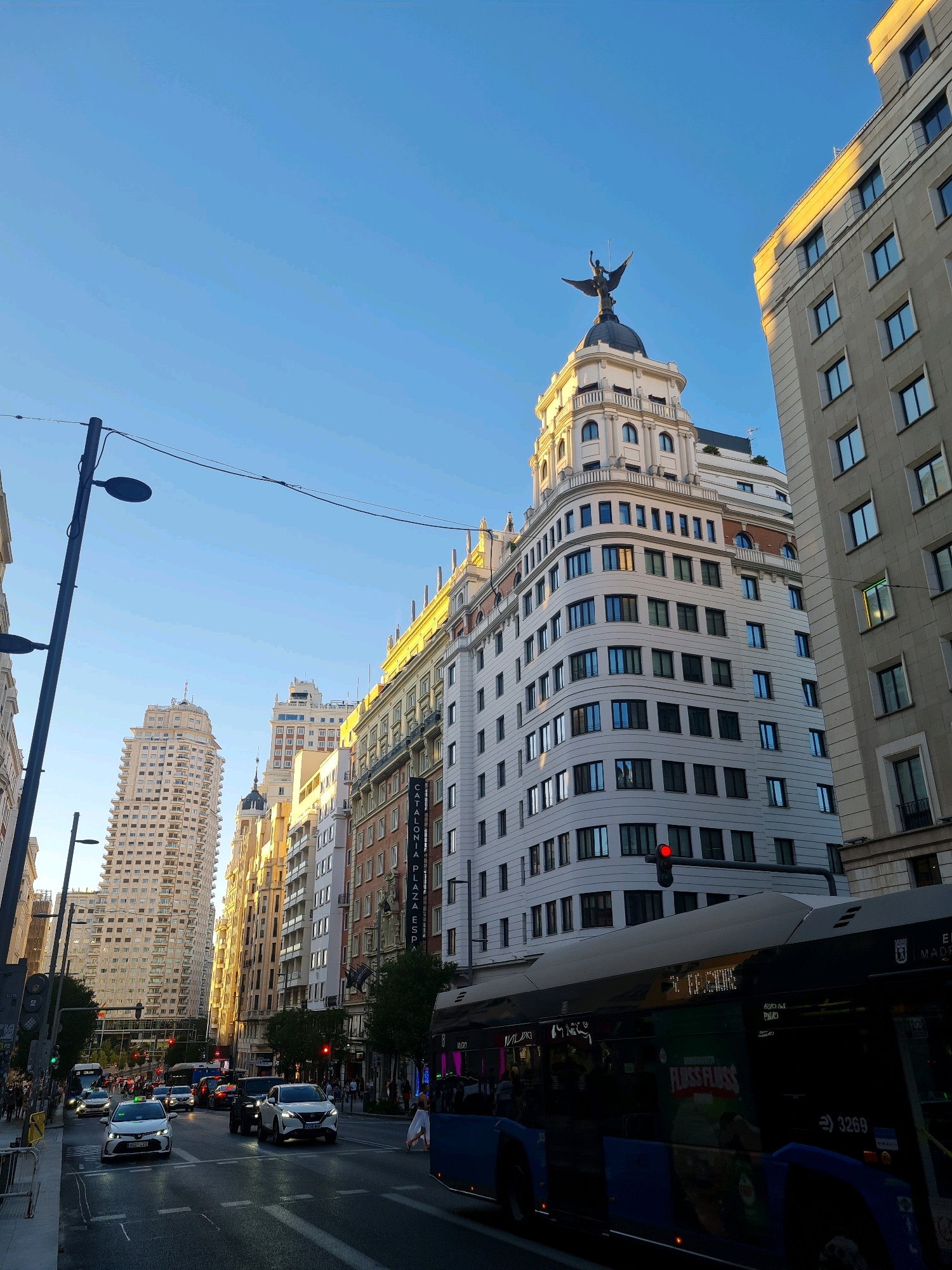 The image captures a view of Gran Vía, a prominent street in Madrid, Spain, bathed in the warm light of late afternoon or early morning. The sky is a clear, bright blue, with a few wisps of clouds visible. Tall, ornate buildings line the street, their facades displaying a mix of architectural styles, from classical to Art Deco.
Dominating the skyline is the Metropolis Building, identifiable by its distinctive dome and the winged figure of Victoria perched atop it. Adjacent to it, to the right, is another imposing building with a grid of windows. Further down Gran Vía, a cluster of tall, pale buildings forms a striking backdrop.
In the foreground, a busy street scene unfolds. A blue public bus, displaying advertisements and the number 3269, is prominent, partially obscuring the view of the road. Several cars and taxis navigate the intersection, with a white taxi visible in the left lane. Traffic lights are visible, with one red light illuminated. Pedestrians can be seen crossing the street and walking on the sidewalks. Streetlights and power lines crisscross the urban landscape. The overall impression is of a vibrant, bustling city street in Madrid.