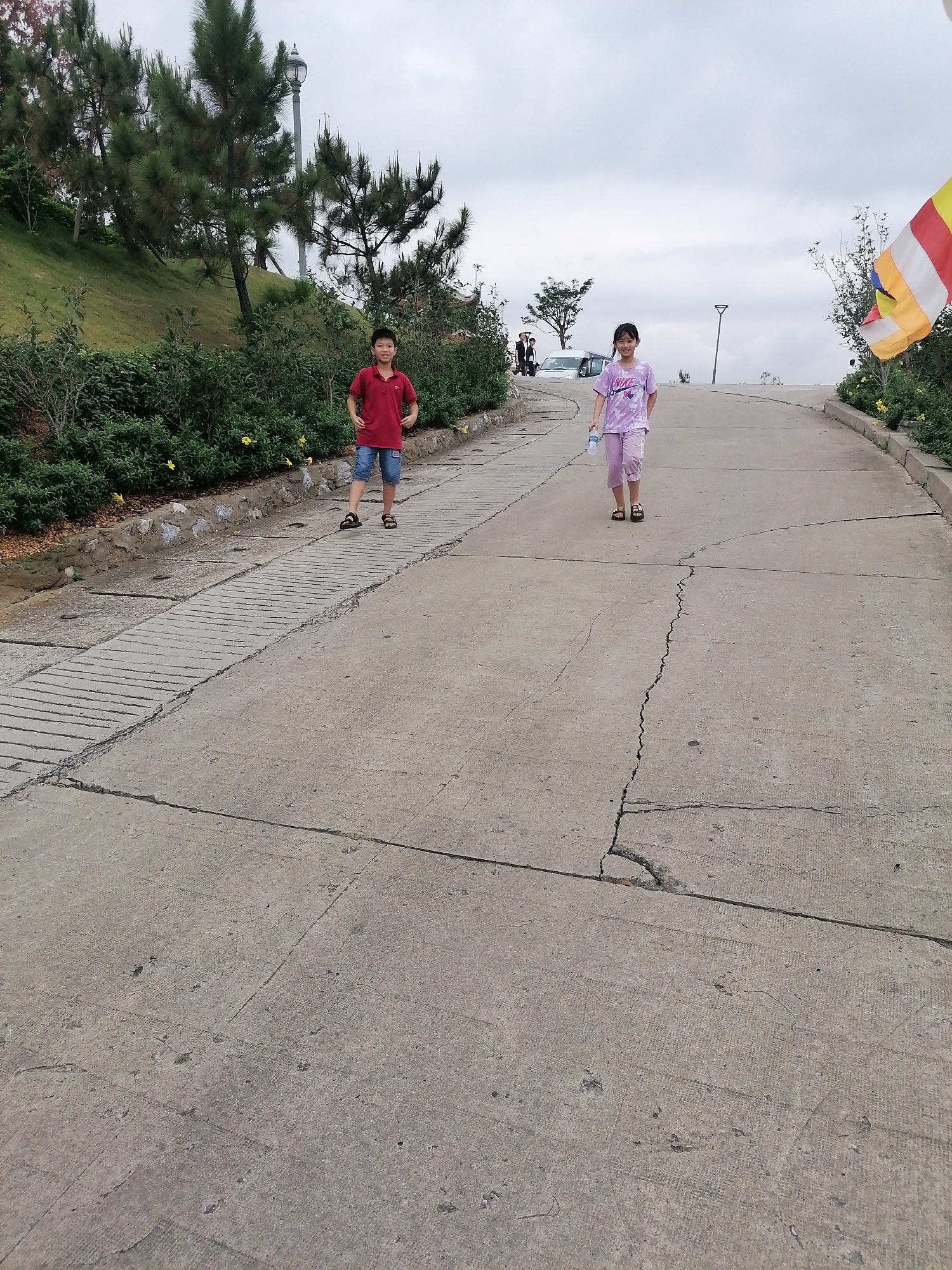 The image captures an outdoor scene featuring two children on a paved, sloping road under an overcast sky. The setting appears to be a hilly area with lush greenery, possibly in Hong Gai Ward, Vietnam, as indicated by the context.
On the left side of the road, a boy stands facing the camera with a slight smile. He is wearing a red short-sleeved polo shirt, blue denim shorts, and dark sandals. Behind him, a green hillside is densely covered with trees, primarily pines, and various bushes. A streetlamp is visible among the trees on the left. The edge of the road features a concrete border and more green shrubs, some adorned with small yellow flowers.
Further up the road and towards the right, a girl walks towards the viewer, also with a gentle smile. She is dressed in a light purple tie-dye t-shirt that displays the brand name "NIKE" on the front, paired with matching purple pants and dark sandals. She holds a clear plastic water bottle in her right hand. In the distance behind her, a white car and another individual can be seen at the top of the incline. A colorful striped flag, possibly a Buddhist flag, with prominent red, yellow, blue, white, and orange sections, is visible on the far right. Another streetlamp is seen further up the road.
The concrete road surface shows significant signs of wear and tear, with numerous cracks running across its width and length, especially noticeable in the foreground. The weather appears cloudy and muted, suggesting a temperate daytime setting. The children seem relaxed and at ease in this natural environment.