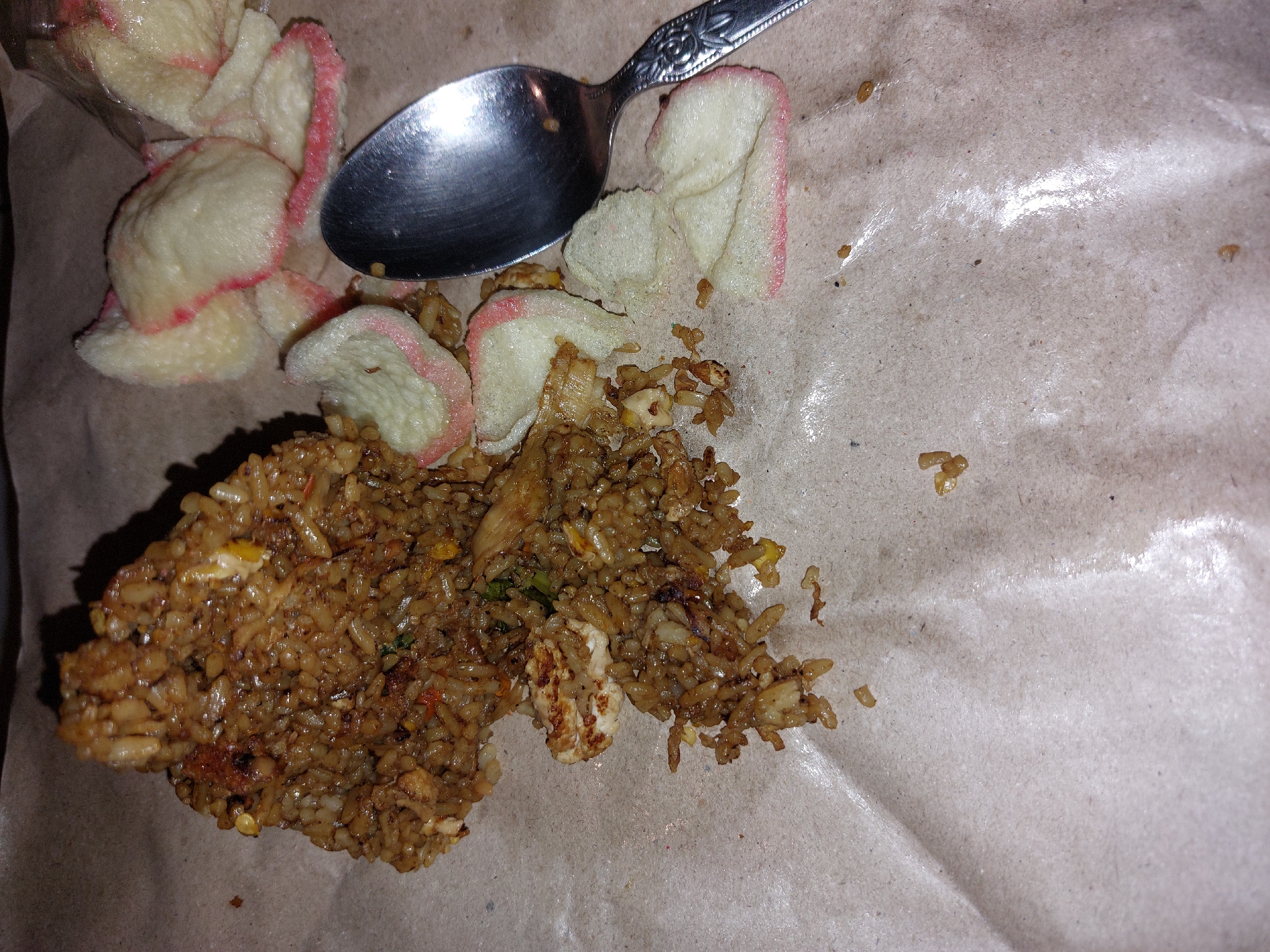 The image shows a close-up, overhead view of a portion of fried rice served on brown paper, likely takeout packaging. A metallic spoon rests on top of the food, its reflective surface catching the light. Scattered around the rice are several light-colored crackers with pink edges, possibly prawn crackers (krupuk). The fried rice appears to be seasoned and contains visible pieces of chicken and other ingredients. The brown paper packaging is slightly creased and shows some oil stains. The lighting is dim, suggesting it might be evening or an indoor setting. There is no visible text in the image. The overall impression is that of a hastily prepared or partially consumed meal, possibly in a casual setting in Karangjati, Indonesia.