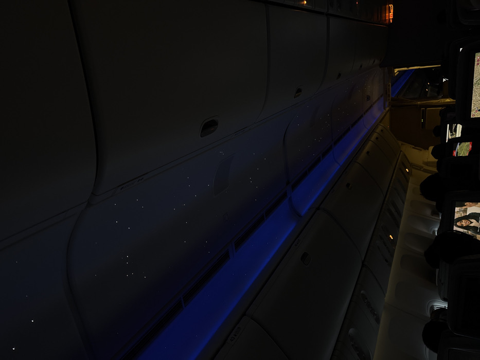 The image displays the interior of a commercial aircraft cabin, observed from a forward-facing perspective, possibly from a passenger seat. The cabin is dimly lit, with a dark ceiling featuring numerous small, white lights arranged to resemble stars, complemented by parallel blue LED accent light strips running lengthwise along the overhead panels. Overhead storage bins are visible along the upper sides of the cabin.
In the foreground and midground, the backs of several dark passenger seats are visible. Each seat is equipped with a personal in-flight entertainment (IFE) screen. One IFE screen in the immediate foreground prominently displays a video of a woman with dark hair, shown from the chest up. Another IFE screen further back on the right side shows a geographical map display. Several passengers are present, their heads and upper torsos appearing as dark silhouettes in their seats. Towards the rear of the cabin, an illuminated orange "EXIT" sign is partially visible above a doorway.
The scene is consistent with an aircraft cabin environment, likely either during nighttime or with dimmed cabin lighting. If this scene is located in Dum Dum, India, it depicts an aircraft at Netaji Subhas Chandra Bose International Airport.