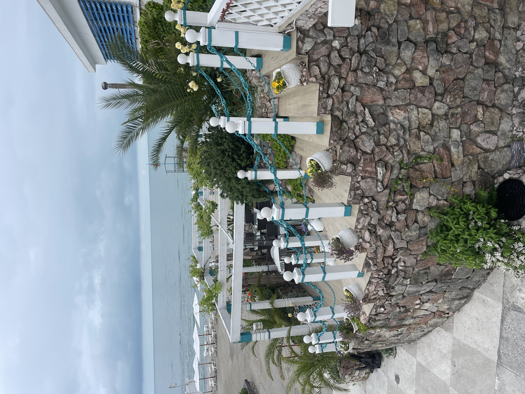 The image shows a coastal scene in Sochi, Russia. A set of stairs leads from a stone wall, adorned with greenery, down to a restaurant or patio area by the water. The stairs have white railings with teal accents and are decorated with potted plants. The restaurant area features white umbrellas and seating. The sea is visible in the background under a cloudy sky. Palm trees are also visible.