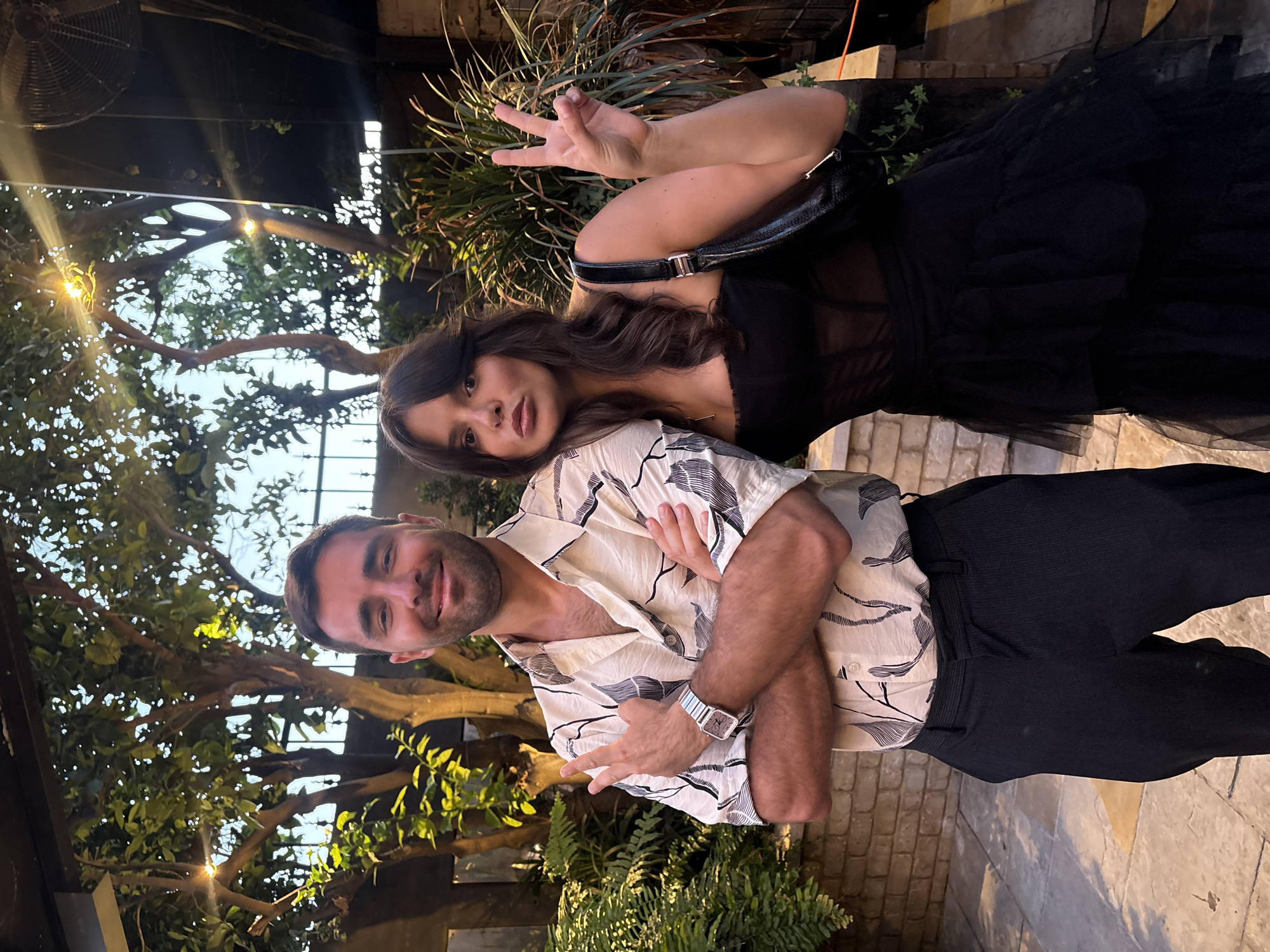 The image shows two people outdoors at night. A man with dark hair and a beard is wearing a patterned shirt, dark pants, and a watch. He has his arms crossed and is making a peace sign. A woman with long dark hair is wearing a black dress and a black purse. She is also making a peace sign. They are standing in front of a brick wall and some greenery. There are trees and lights in the background. The location cannot be determined to be in the city of Tel-Aviv, Israel.