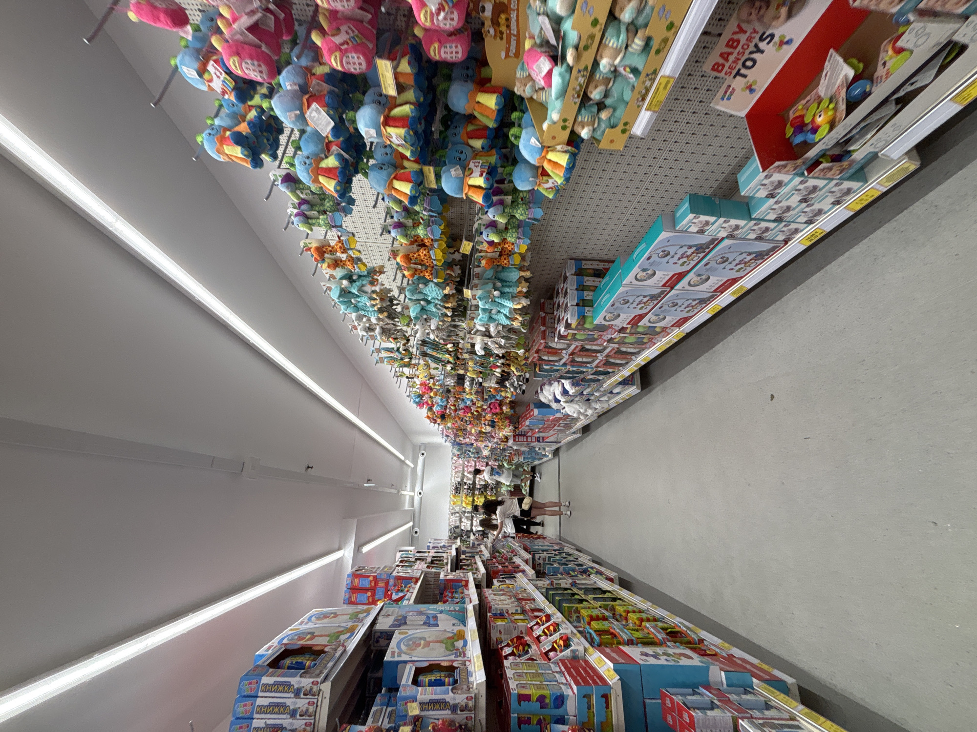 The image displays an interior view of a well-lit toy store aisle located in Plovdiv, Bulgaria. The perspective is down the center of the aisle, which features extensive white metal shelving units on both sides. The floor is a uniform light gray. Overhead, multiple long, rectangular fluorescent light fixtures illuminate the space.
On the left side of the aisle, the shelves are primarily stocked with boxed items, likely children's games or educational materials, with the Cyrillic word "КНИЖКА" (KNIZHKA), meaning "booklet" or "small book," visible on some product packaging.
The right side of the aisle presents a dense display of colorful merchandise. The upper sections feature numerous small plush toys, including blue octopuses, various animal figures (such as orange and green giraffes), and pink items resembling toy phones, all hanging from hooks on perforated back panels. Below these hanging items, boxed baby mobiles are neatly stacked, and further down, open displays contain assorted colorful plastic baby toys, some labeled "BABY SENSORY TOYS" in English.
Approximately four individuals are visible in the mid-ground of the aisle, all appearing to be adults or young adults. One woman, wearing a white top and dark shorts with white sneakers, stands in the foreground, facing the right-side products. Behind her, three other patrons are discernible, including a man in a white t-shirt and dark shorts who is also observing items on the right-side shelves. The individuals are engaged in browsing the merchandise. The store appears clean, organized, and well-maintained.