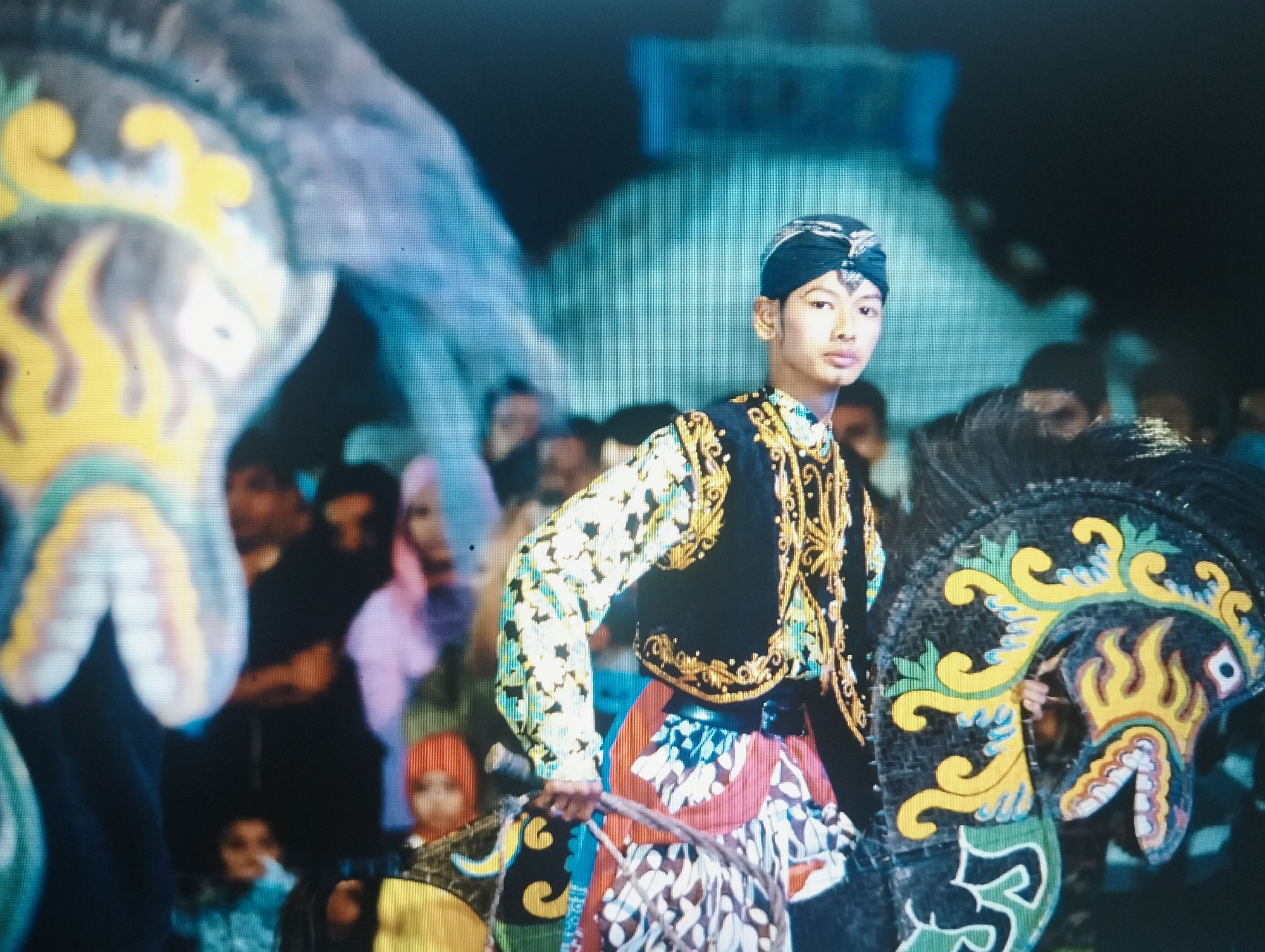 The image depicts a scene at night, possibly a cultural performance or festival, in the City of Medan, Indonesia. In the foreground, a young man dressed in traditional attire stands out. He wears a black turban with silver ornamentation, a black vest adorned with gold embroidery, and a brightly colored patterned shirt and skirt. He is holding onto what appears to be a prop, possibly part of a costume or a puppet, which has a stylized, colorful face with a red mouth and yellow flames. To the left of the young man, another large, brightly colored prop, possibly an animal's head or a float, is visible, featuring yellow flames and a blueish-gray body. In the background, several other individuals are present, some blurred and out of focus, indicating a crowd. The environment appears to be illuminated by artificial lights, with a dark background that suggests nighttime. The overall atmosphere suggests a celebratory or ceremonial event. The style of clothing and the presence of elaborate props are indicative of Indonesian cultural traditions, possibly a wayang or similar performance.