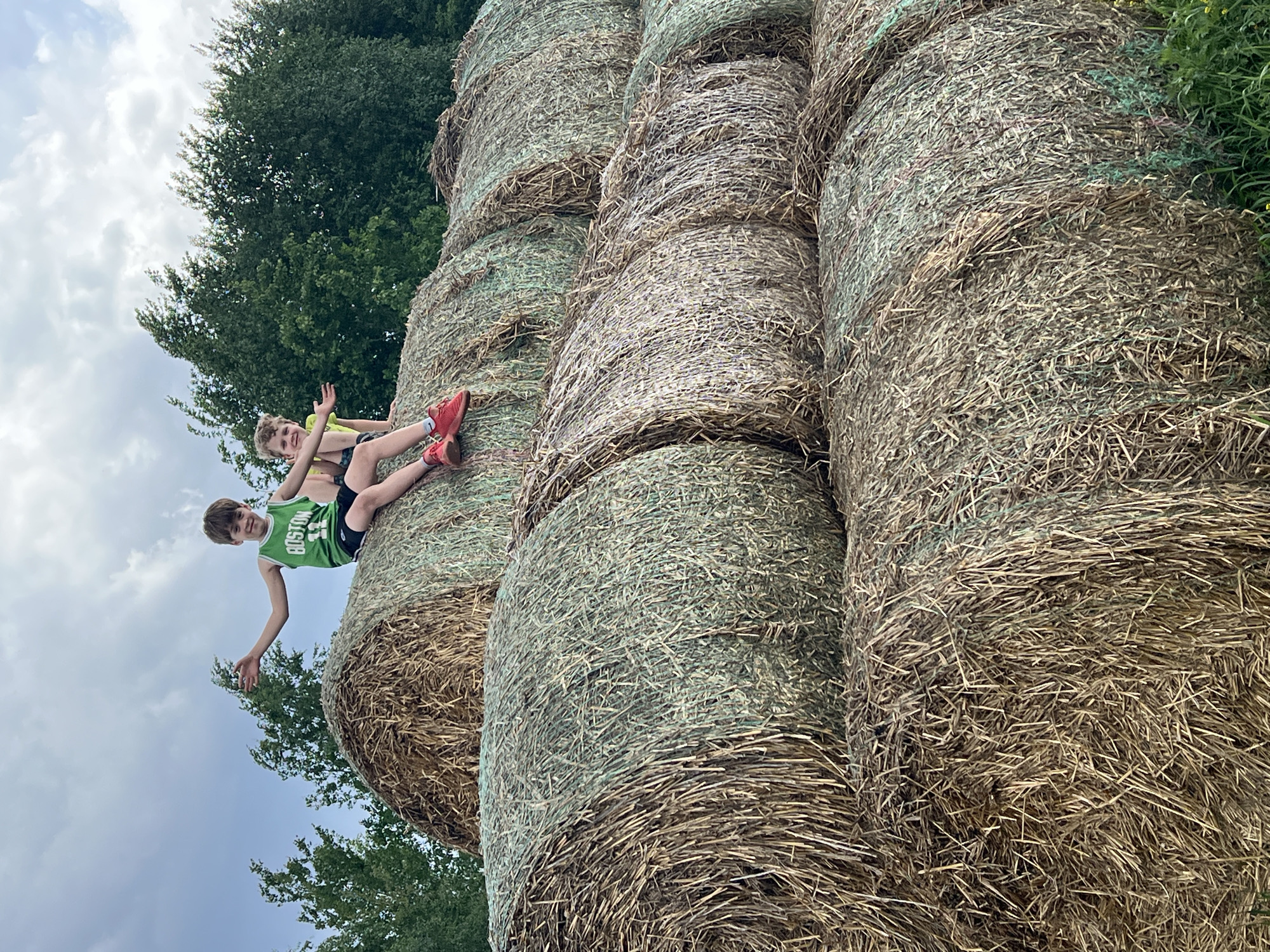 Two children sit atop stacked hay bales outdoors. One child wears a green "Boston" jersey and black shorts. The other child is in a yellow shirt and patterned shorts, wearing red sneakers. Behind the hay bales are trees, and the sky is partly cloudy. There is no information indicating the location is Kirillovka, Russia.
