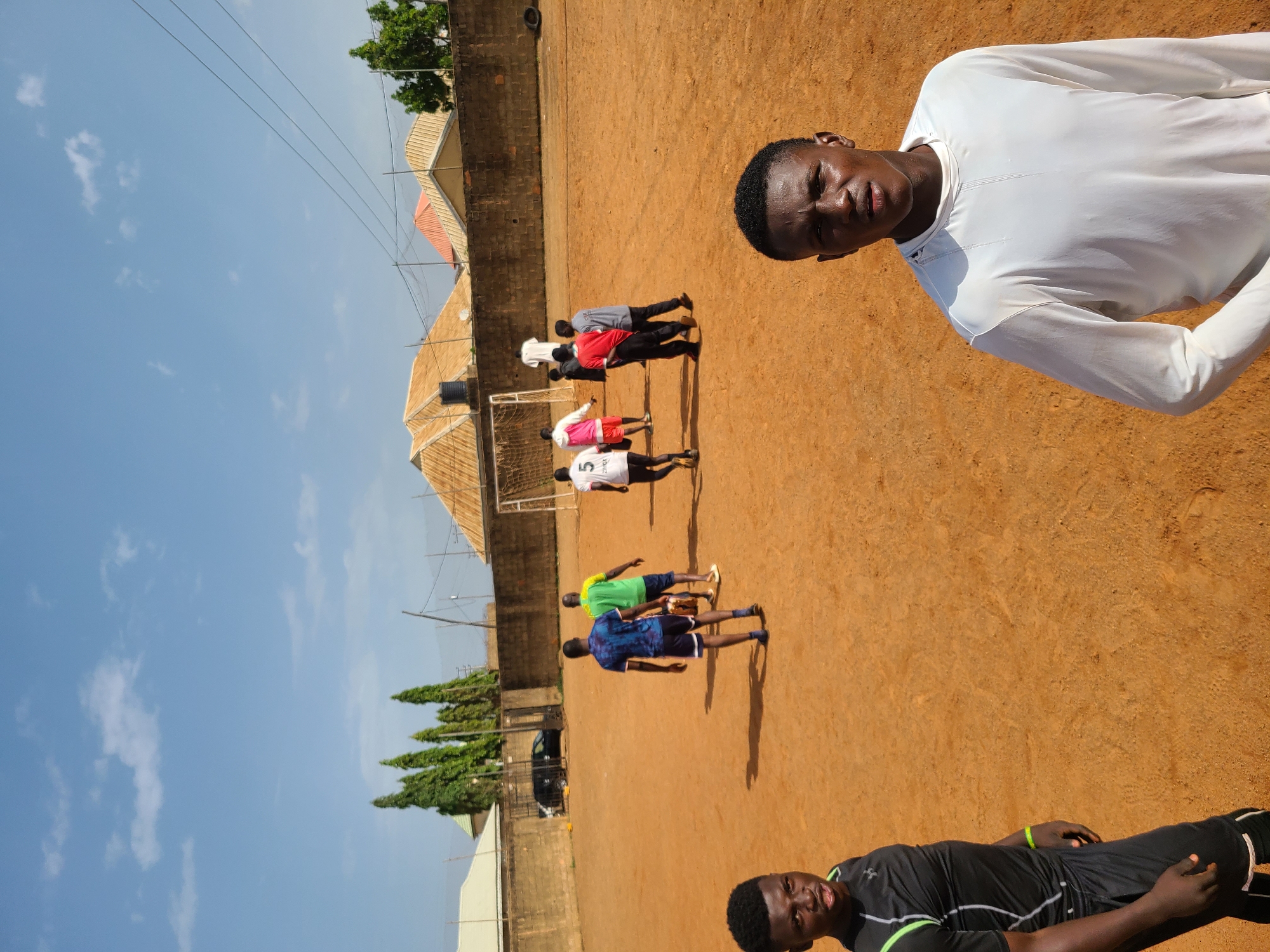 The image captures a group of young men on a dirt soccer field under a clear blue sky, suggesting a sunny day. The scene appears to be in Barakin Lerawi, Nigeria, indicated by the context provided. Several individuals are present on the field, some actively engaged in what seems to be a soccer practice or game.
In the foreground, a young man in a white long-sleeved shirt is positioned close to the camera, looking directly at it with a determined expression. His skin is glistening with sweat, hinting at physical exertion. Further back, a line of young men are spread across the field. One is wearing a white jersey with the number 5, another a red shirt, and others are in blue and green tops. They appear to be in various stances, possibly during drills or a match. A soccer goal is visible in the background on the left side of the image.
The setting includes a low brick wall bordering the field and buildings with corrugated iron roofs visible behind it, typical of some residential areas in Nigeria. Some trees are also present, adding to the landscape. The overall atmosphere seems to be one of active recreation and youth engagement in sports. The lighting and shadows suggest it's daytime.