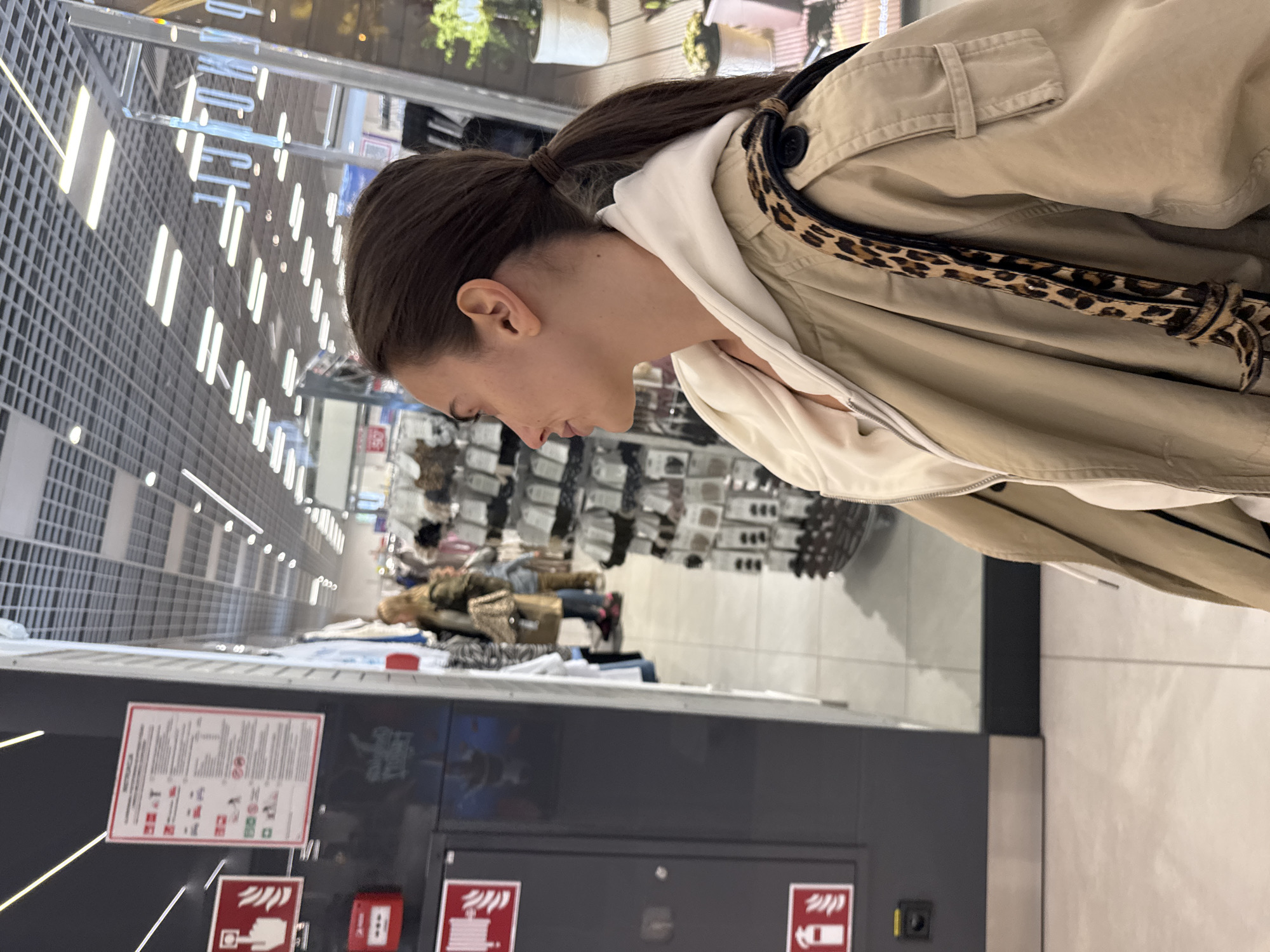 The image captures a candid moment inside what appears to be a retail store or department store, likely in Warsaw, Poland, based on the visible Polish text. The primary subject is a woman, seen from the side in a medium shot, looking downwards with a focused, neutral expression. She has dark hair styled in a high ponytail. She is dressed in a light beige or tan jacket, worn over a white hooded top. A distinct leopard print strap or belt is visible over her shoulder.
The background is a bustling but somewhat blurred indoor commercial environment. A prominent feature of the setting is the modern ceiling, characterized by a grid-like metallic structure interspersed with linear artificial lights, creating bright illumination. Shelves and display racks laden with various clothing items or merchandise can be faintly discerned further back, with other shoppers visible in the distance, out of focus.
In the lower left foreground, a dark grey panel houses official-looking signs. A rectangular white sign with red lettering and pictograms is clearly visible, with the Polish word "INSTRUKCJA" (Instruction) prominently displayed at the top, indicating it's likely a fire safety or emergency procedure notice. Below this, two red square symbols with white icons (resembling a hand pressing a button and a fire symbol) are positioned, typical of fire alarm call points or safety equipment indicators. The woman's activity suggests she is browsing or observing items within the store. The overall impression is a casual, daytime scene in a public commercial space.