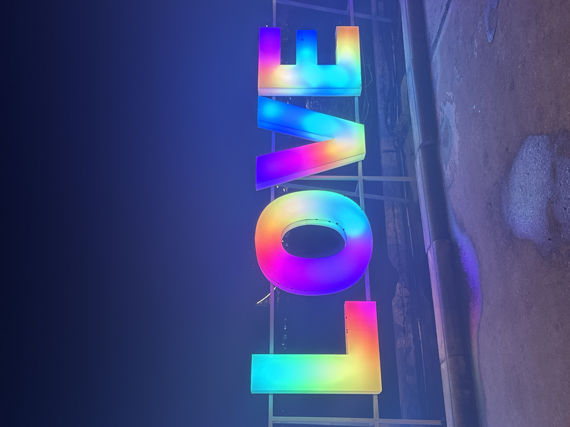 The image features a large, horizontally aligned illuminated sign spelling the word "LOVE" against a dark blue night sky. Each letter is independently lit with a dynamic rainbow color gradient, with hues transitioning vertically across their surfaces. The sign is positioned behind a light-colored metal railing. In the distant background, faint lights are discernible on the horizon, and a slender, illuminated structure is visible between the letters 'L' and 'O'. The foreground consists of a dark, textured concrete or paved surface with wet patches, reflecting the vibrant rainbow colors of the sign. A low, dark concrete curb runs along the base of the railing. The scene is captured at night in ตำบลเกาะพะงัน, Thailand.