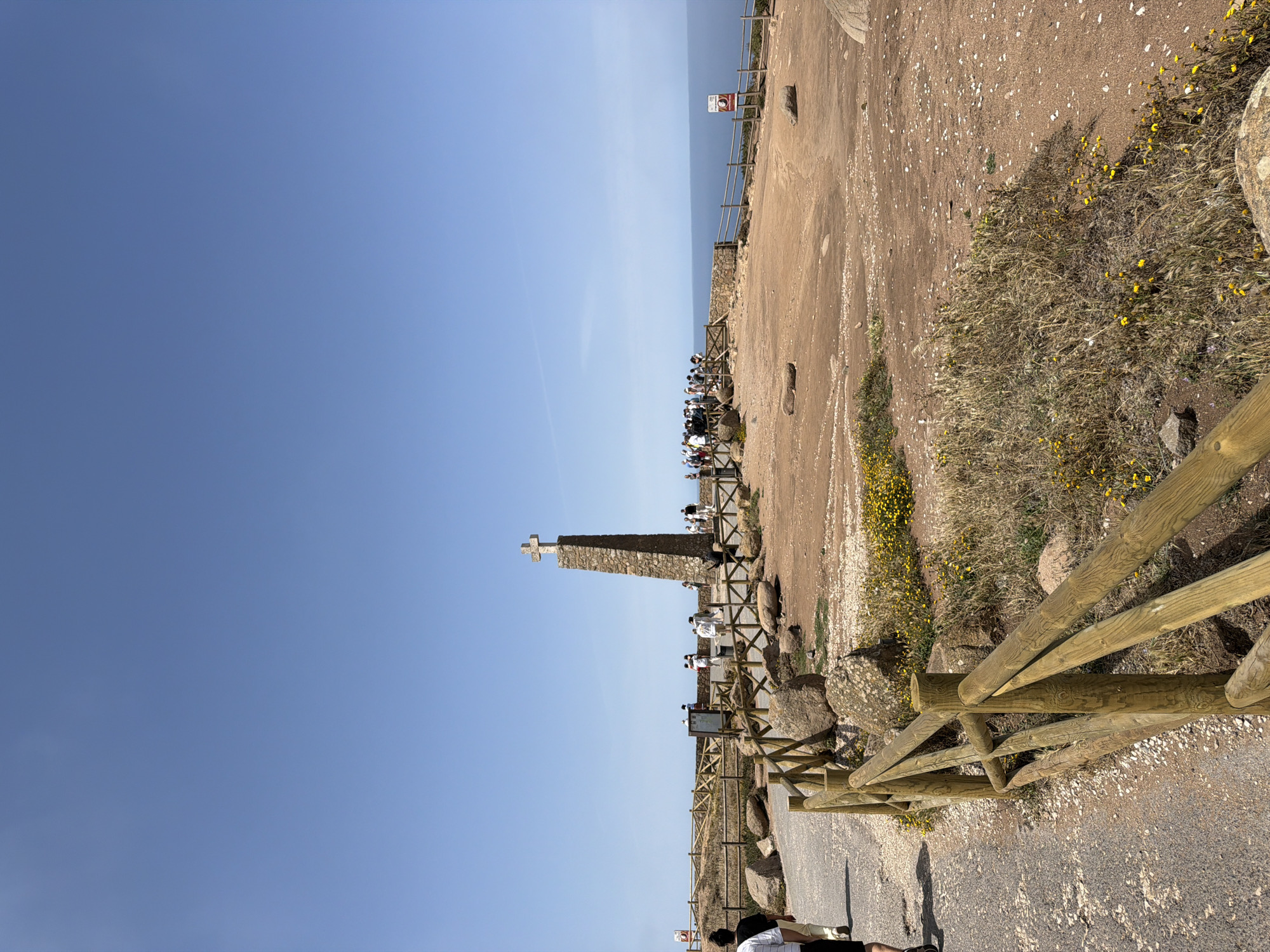 The image depicts Cabo da Roca, the westernmost point of mainland Europe, located near Colares, Portugal. A stone monument with a cross stands prominently in the center, surrounded by a group of tourists. The landscape is characterized by dry, reddish soil, scattered rocks, and sparse vegetation with yellow wildflowers. A wooden fence demarcates a pathway leading towards the monument and a viewing area overlooking the vast blue ocean. The sky is clear and bright blue. Several people are visible walking and observing the scenery, with some gathered around the monument and others near the edge of the cliff. The overall scene suggests a popular tourist destination on a sunny day.