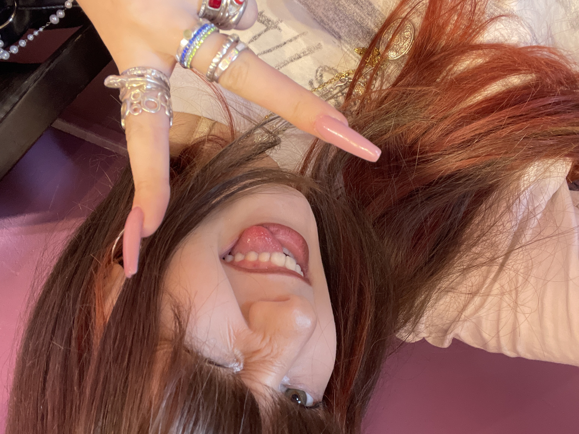 A woman with brown and red hair is lying on a pink surface, her head tilted towards the camera. She sticks her tongue out and winks one eye. She has multiple rings on her fingers, including a large silver ring and a colorful ring with blue and green stones. Part of her left shoulder and a white shirt are visible in the background. The environment indicates this is a personal photograph, most likely taken indoors. There is no information to conclude that this image was taken in Raanana, Israel.