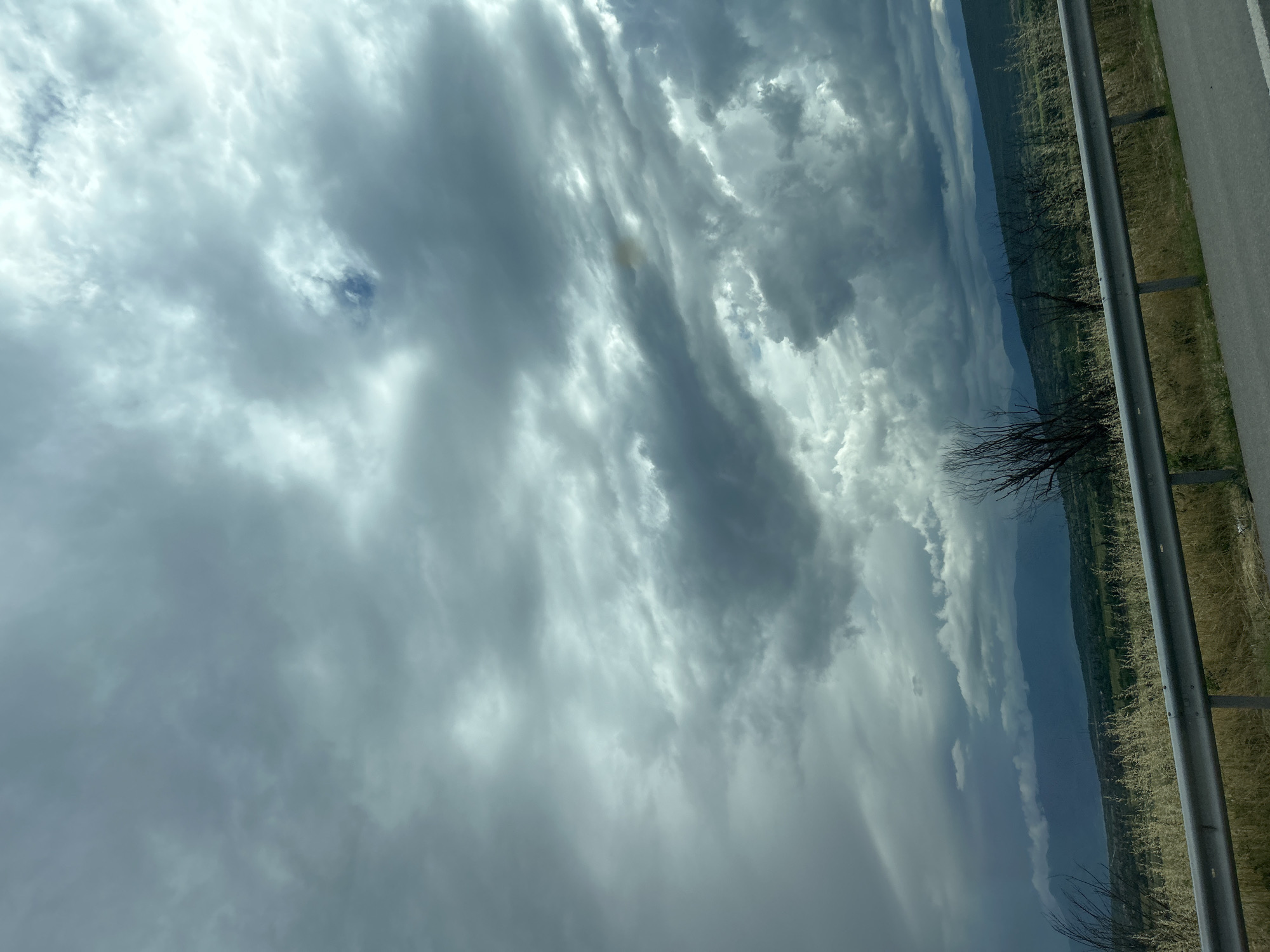 The image captures a dramatic cloudy sky above a rural landscape, viewed from the side of a road in Kalugerovo, Bulgaria. The sky is largely covered by a vast array of clouds, displaying varying shades from bright, luminous white to dark, heavy grey formations, with small patches of blue sky visible in some areas. This indicates an overcast or potentially pre-storm weather condition during daylight hours.
Below the expansive and dynamic cloud cover, a distant panorama of rolling hills or mountains stretches across the mid-ground, appearing verdant but shadowed, contributing to the atmospheric mood. In the immediate foreground, on the right side of the frame, a robust metal guardrail runs horizontally, separating a strip of dry, light-colored grass and scattered, bare tree branches from a narrow edge of an asphalt road. No human activity or visible text is present, emphasizing the natural landscape and the overwhelming presence of the sky.