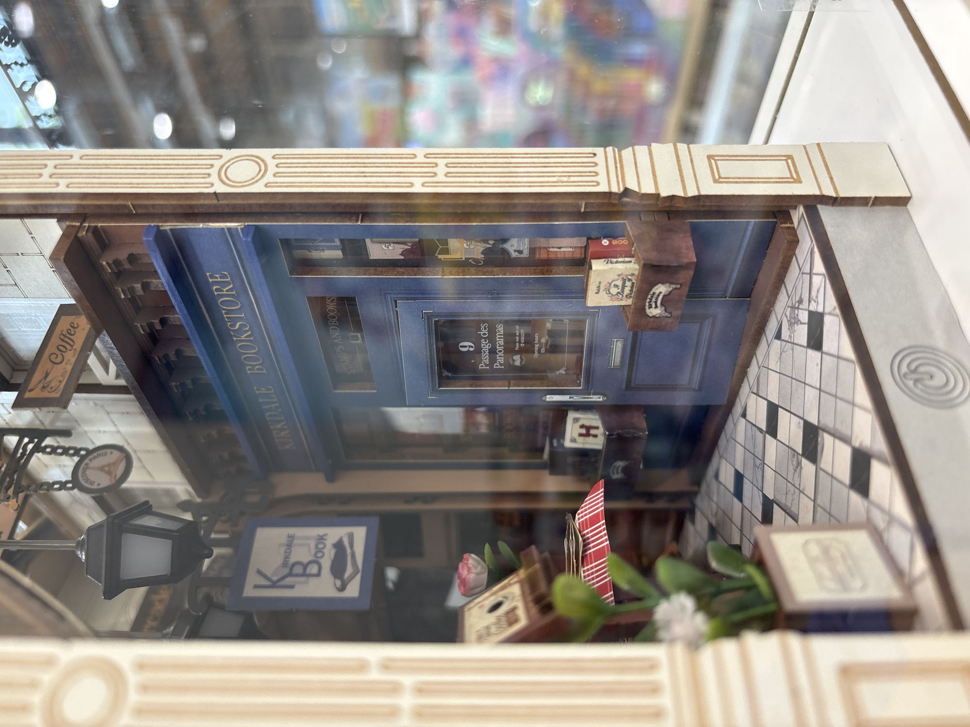 The image shows a miniature diorama of a bookstore, titled "Kirkdale Bookstore." The diorama is enclosed, possibly in glass or plastic, as evidenced by reflections on the surface. The scene depicts the storefront of the bookstore, with a blue facade and signage that reads "Kirkdale Bookstore, Stamps and Books." The door of the shop is labeled "9 Passage des Panoramas," and includes opening hours from 10am to 6pm.
Additional elements include a "Coffee" sign, a street lamp, and a smaller sign indicating "Kirkdale Book" with a cat silhouette. Books and decorative items are visible inside and outside the store. A tile pattern is visible along the right side of the scene. The diorama features a frame with patterned edging. The diorama appears to be inside, with a reflected background of an indistinct indoor location.
The city of Choeng Thale, Thailand, is not visibly represented or implied in the image.