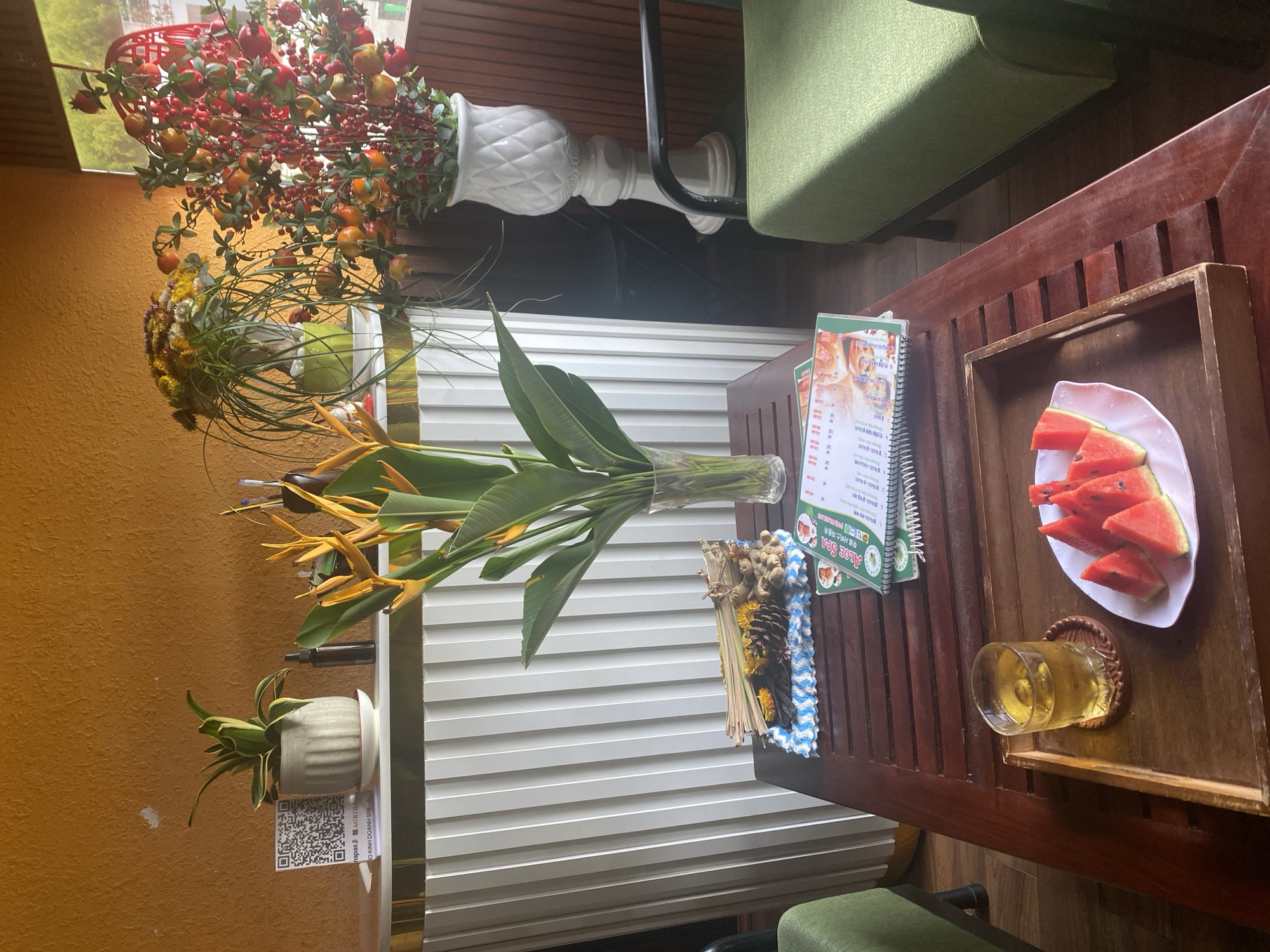 In Mũi Né, Vietnam, a low wooden table sits in a brightly lit outdoor setting, possibly a cafe or reception area. On the table, a wooden tray holds a glass of amber liquid filled with ice and a white plate with several slices of watermelon. To the left of the tray, a decorative arrangement of dried reeds, pinecones, and small yellow flowers rests on a blue and white patterned cloth.
Behind the table, a counter with a white, ribbed facade extends across the frame. On the counter, a QR code is displayed on a white card next to a potted green plant. To the right of the plant, a dark spray bottle and a vase containing bright orange heliconia flowers are visible. Further to the right, two decorative plants with red berries and orange fruits in white vases are positioned, one tall and ornate, the other shorter.
Two green chairs are partially visible on either side of the scene. The wall behind the counter is a textured ochre color, and a wooden paneled wall forms the background to the right, where a window or open doorway reveals a glimpse of greenery. A spiral-bound booklet, likely a menu, lies open on the table, displaying text and images. The overall atmosphere is relaxed and inviting.