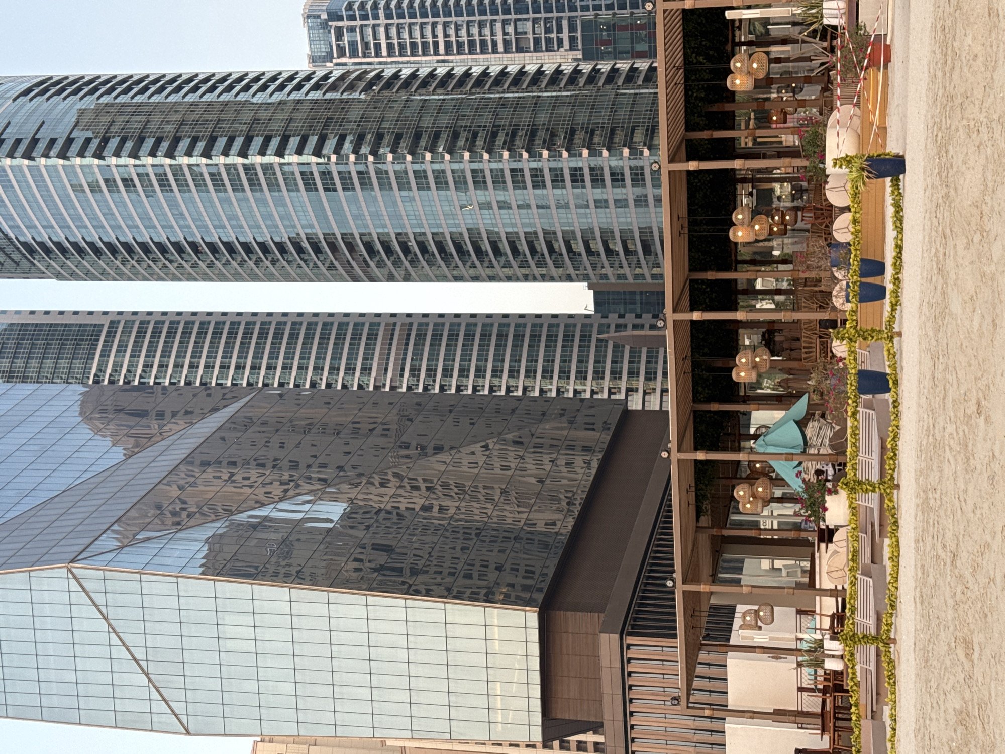 The image shows a beachfront cafe with a view of modern skyscrapers in Dubai, United Arab Emirates. The cafe has a wooden structure with a thatched roof, adorned with woven pendant lamps. It is furnished with tables, chairs, and lounge chairs, suggesting a relaxed atmosphere. A turquoise umbrella provides shade. The foreground is a sandy beach with a low decorative fence made of green foliage and yellow flowers. In the background, several tall glass skyscrapers dominate the skyline, reflecting the sky and surrounding buildings. One building has a distinctive angular glass facade, while others are cylindrical with rows of balconies. The overall scene depicts a harmonious blend of natural beach elements and urban architecture, characteristic of Dubai's coastal developments. No people are visible in the image.