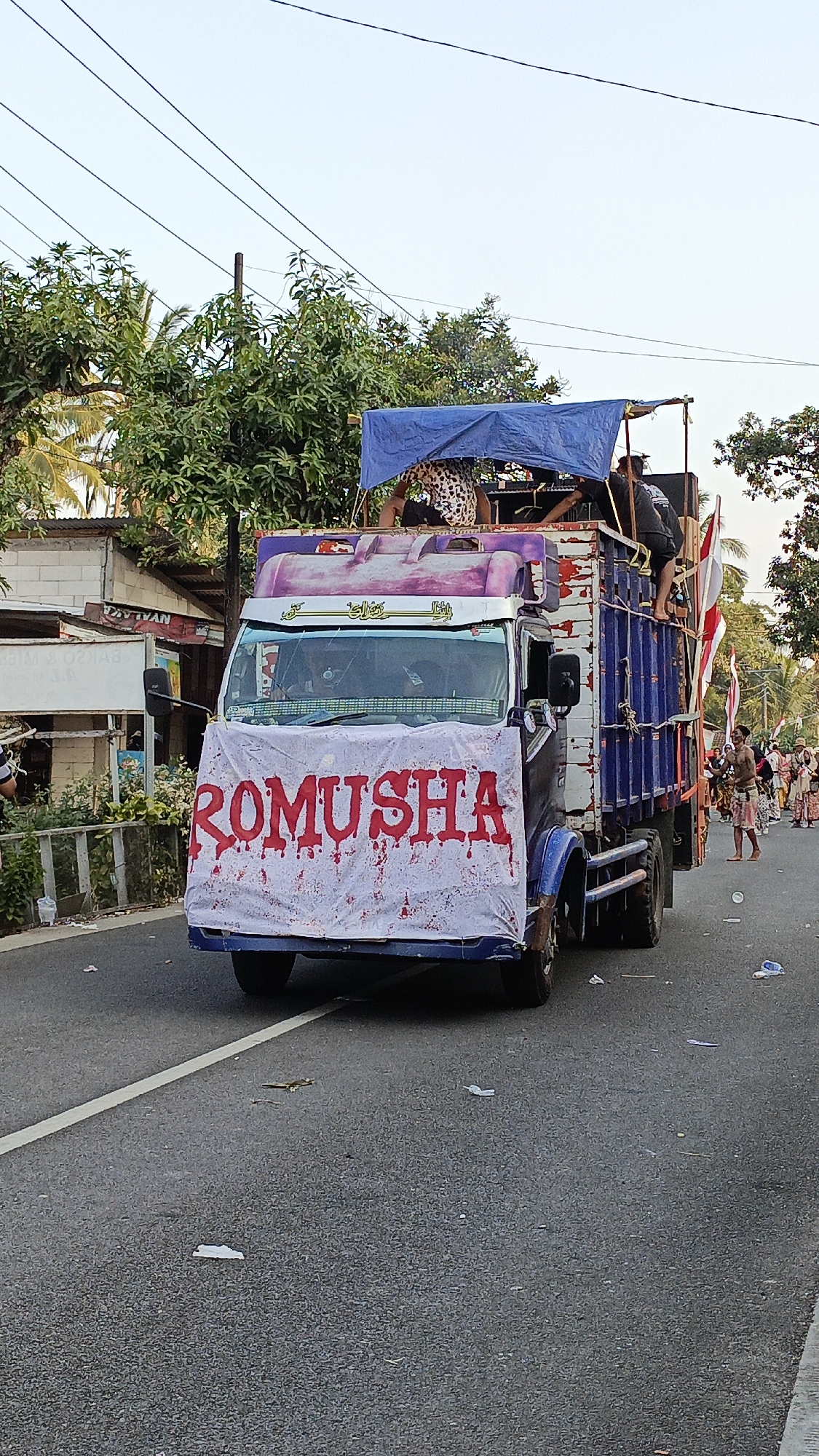 A truck is driving down a paved road in Krajan Lor, Indonesia. The truck is decorated with a banner that reads "ROMUSHA" in red, bloody-looking letters. A blue tarp is draped over the top of the truck, and several people are standing and sitting on top of the truck. There are speakers and other equipment on the truck as well. The truck is adorned with Indonesian flags. People are walking on the side of the road. The surroundings include trees and buildings. The sky is clear and a pale blue. The road has a white line marking the lane. There is trash scattered on the road.