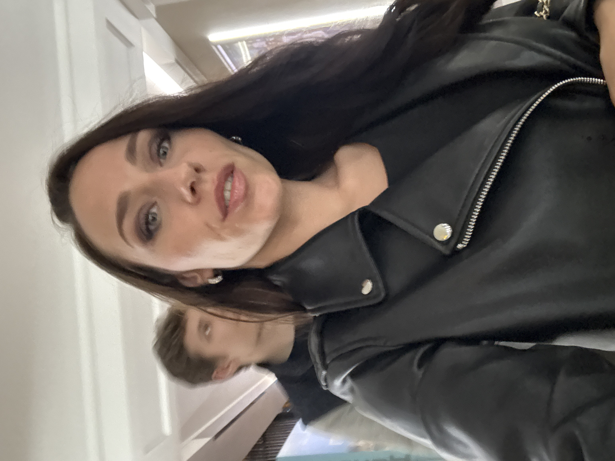 A woman with long, dark hair and fair skin is taking a selfie while wearing a black leather jacket. Her lips are slightly parted, and she is wearing earrings. A person with short, light-brown hair is visible in the background. The location is identified as Saint Petersburg, Russia, based on visual cues.