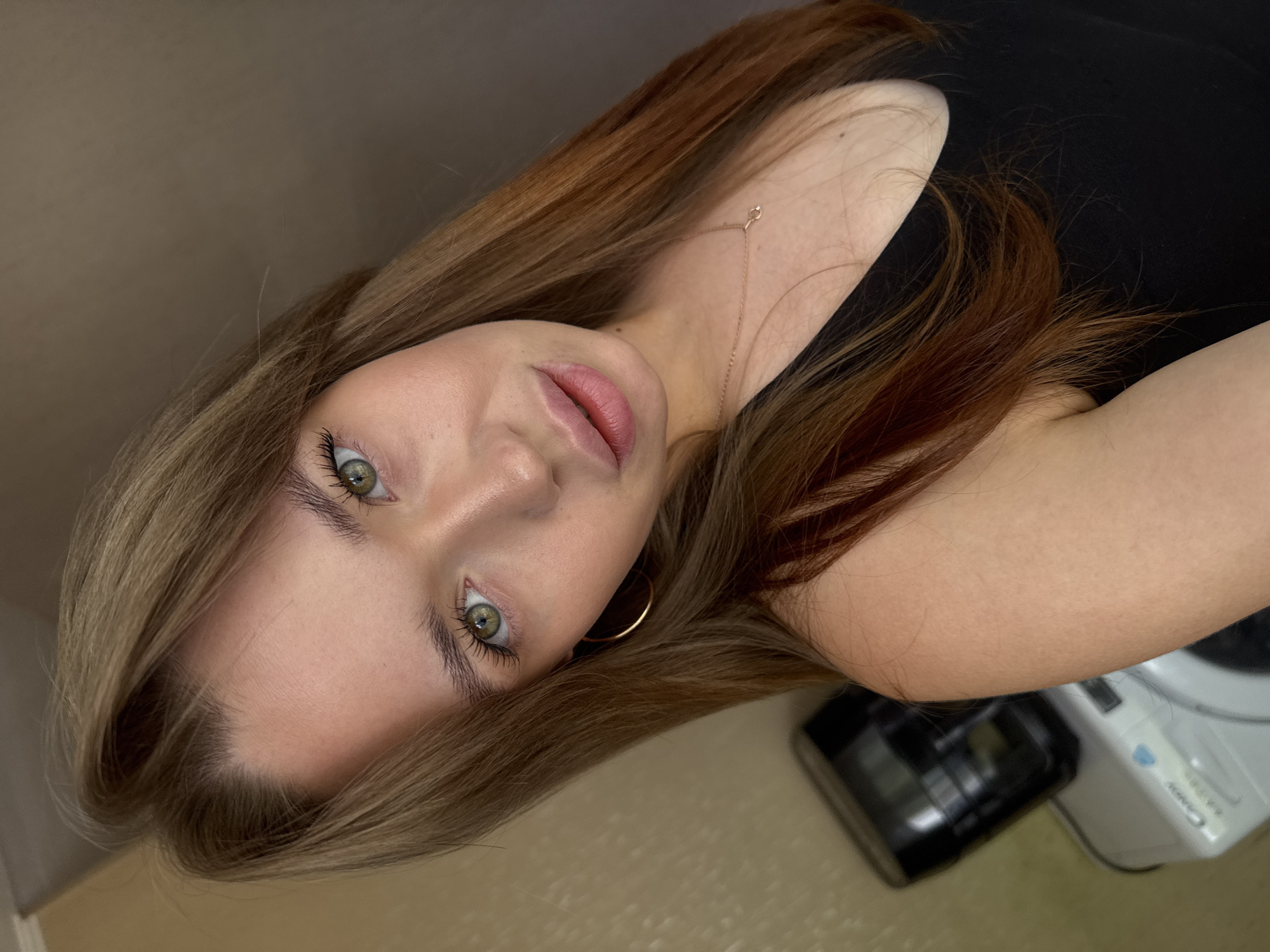 The media file is a close-up, low-angle, portrait-style photo of a young woman. She has long, wavy, light brown and auburn hair that frames her face. Her eyes are a striking green, and she is wearing mascara. Her lips are slightly parted and appear to have a pink tint. She is wearing a black top and a delicate gold necklace. A gold hoop earring is visible in her left ear. The background is out of focus, with a light brown wall or surface on the left and a blurred area with hints of a white appliance and a black object on the right. The lighting appears to be artificial and soft, casting a gentle glow on her face. The overall mood is intimate and contemplative, with the subject looking directly at the camera. No specific time of day or weather conditions are discernible. There is no visible text in the image.