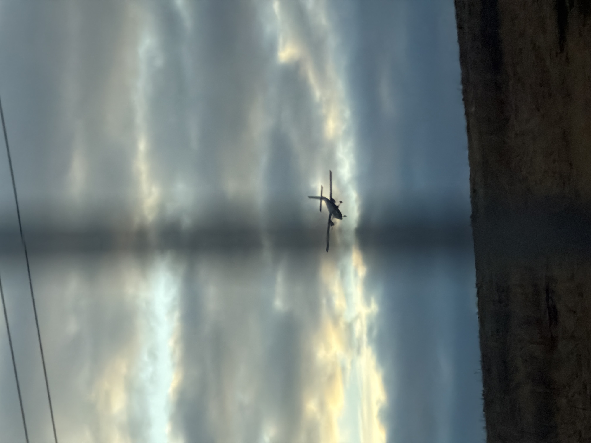 The image depicts a helicopter in flight against a backdrop of cloudy skies and a blurred landscape. It is unclear if this is Yemelyanovo, Russia, or not. Power lines are visible on the left side of the frame. The landscape to the right is dark and indistinct.