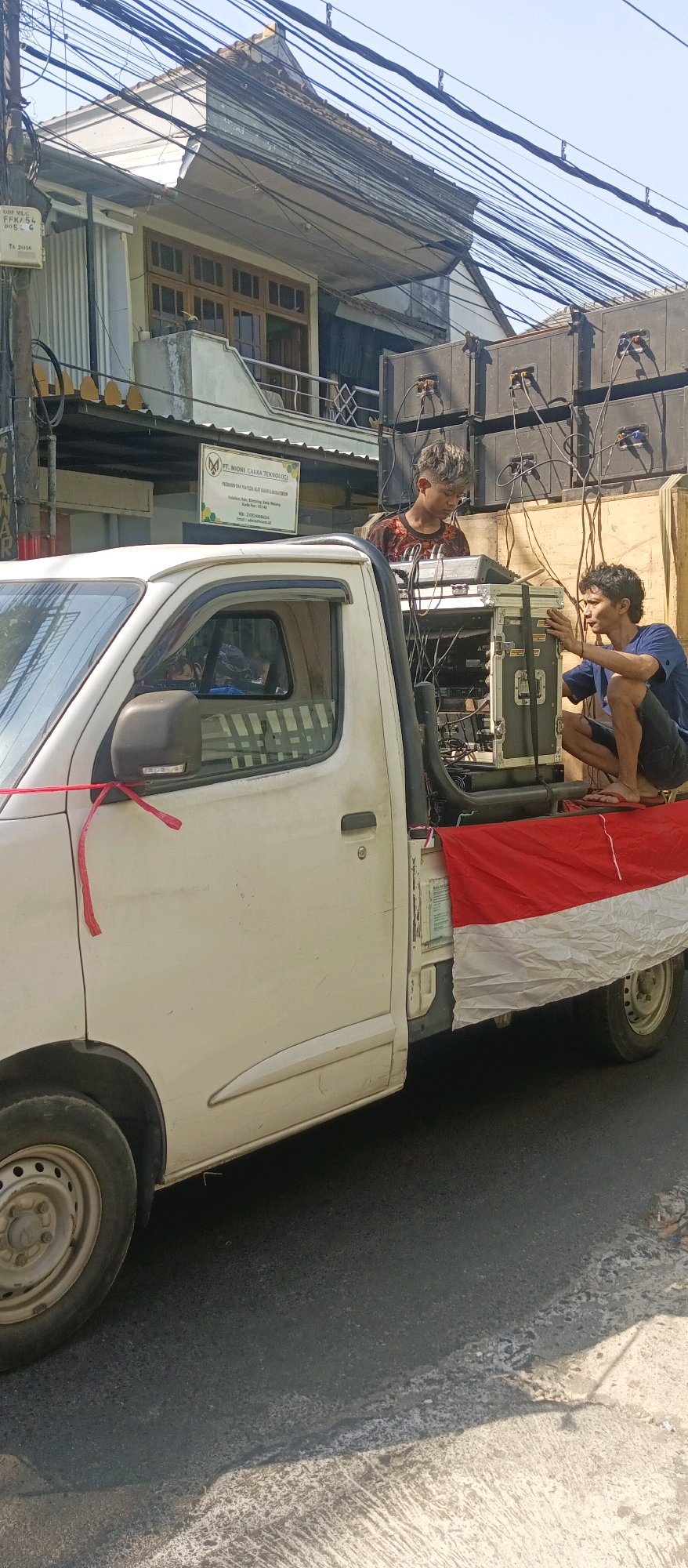 In Malang, Indonesia, a white pickup truck is parked on the side of a street. Two young men are working on audio equipment loaded onto the truck bed. One man, with spiky hair, is operating a mixing board. The other man, with curly hair, is crouching and adjusting equipment. Speakers are stacked behind them. The truck bed is decorated with the Indonesian flag. A sign for "PT. MIONS CAKRA TEKNOLOGI" is visible on a building in the background. The scene takes place outdoors on a sunny day, with overhead power lines and a residential building visible.