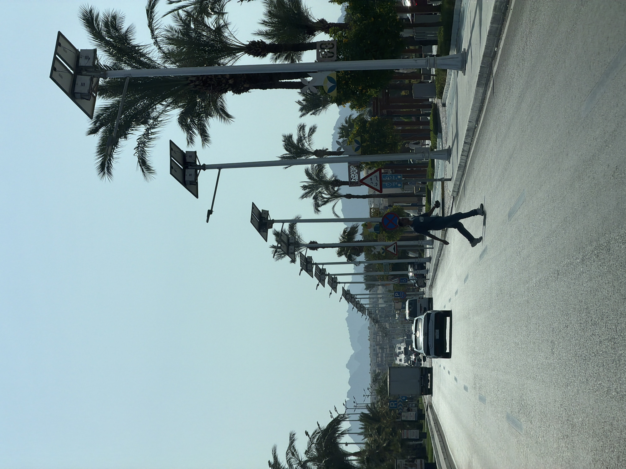 The image captures a daytime view of a wide, paved road in Sharm El Sheikh, Egypt, lined with palm trees and solar-powered streetlights. A person is crossing the road, mid-stride, from right to left. Several vehicles are visible on the road, including a white sedan in the foreground, a white van further ahead, and a truck to the left. In the background, distant mountains are faintly visible against a clear sky. The road appears to be a main thoroughfare, with multiple lanes and clear lane markings. The environment suggests a sunny, potentially warm climate, common to the region.