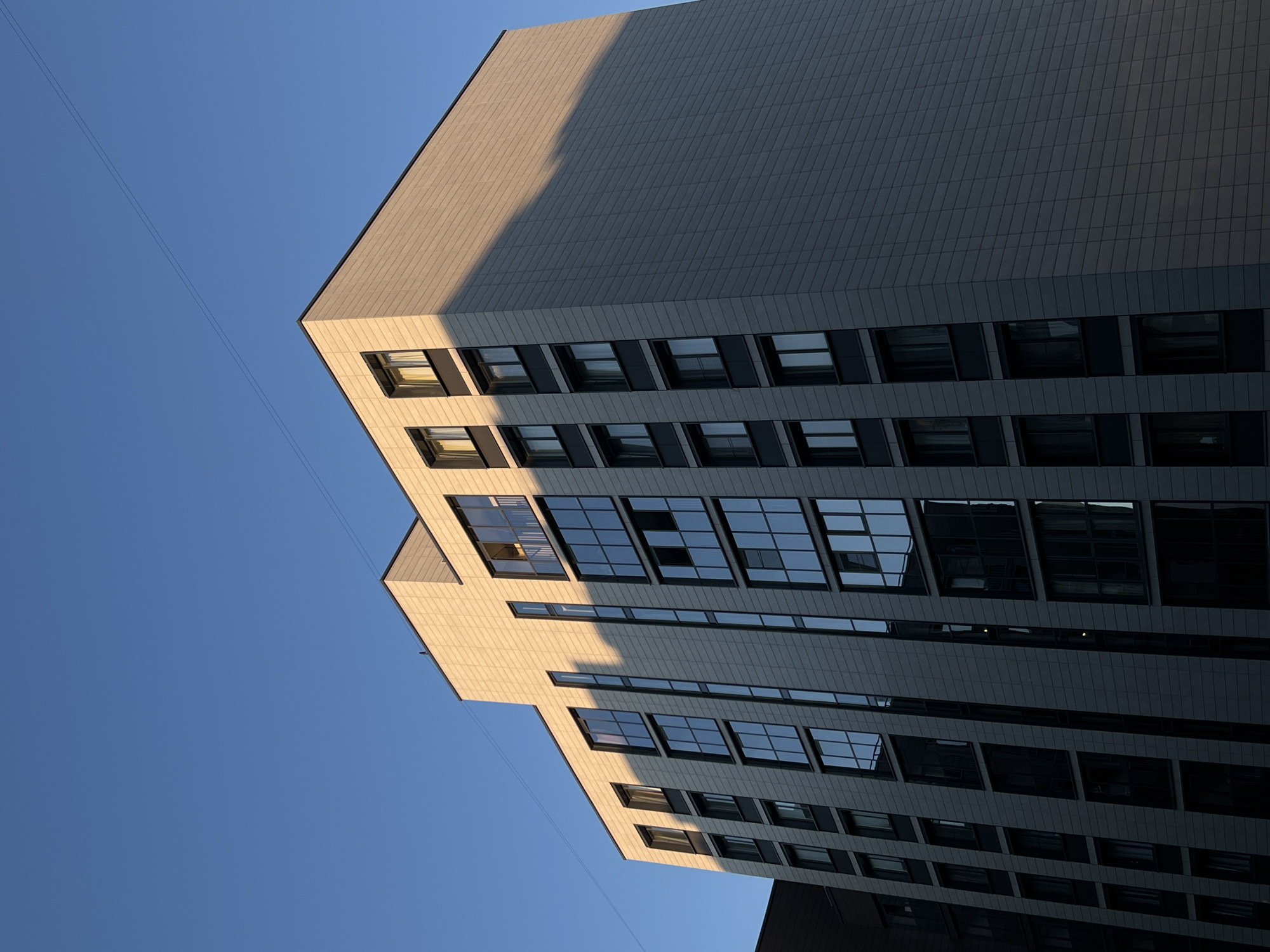 An architectural photograph captures a modern multi-story building from a low, upward-looking angle, rotated to create a dynamic, diagonal composition. The building, likely located in Saint Petersburg, Russia, is made of a grid of light-colored, possibly beige or light grey, rectangular facade panels. A sharp, diagonal shadow line dramatically bisects the building, indicating strong, direct sunlight.
The upper-left and lower-right portions of the building's facade are bathed in warm, golden light, while the central and left-facing sections are cast in deep shadow, appearing dark grey. Numerous rectangular windows are visible across the building's surface. Those in the brightly lit areas reflect the clear blue sky, while windows in the shadowed parts appear darker, some showing glimpses of interiors or muted reflections. A few windows in the lighted section appear to be slightly ajar.
The background consists of a cloudless, vibrant blue sky. Two thin, parallel lines, possibly utility cables or wires, stretch diagonally across the sky in the upper-left and lower-center of the frame. There are no people or discernible activities taking place. The strong, directional lighting and warm tones suggest either early morning or late afternoon (golden hour). The weather is clear and sunny. No visible text is present in the image.